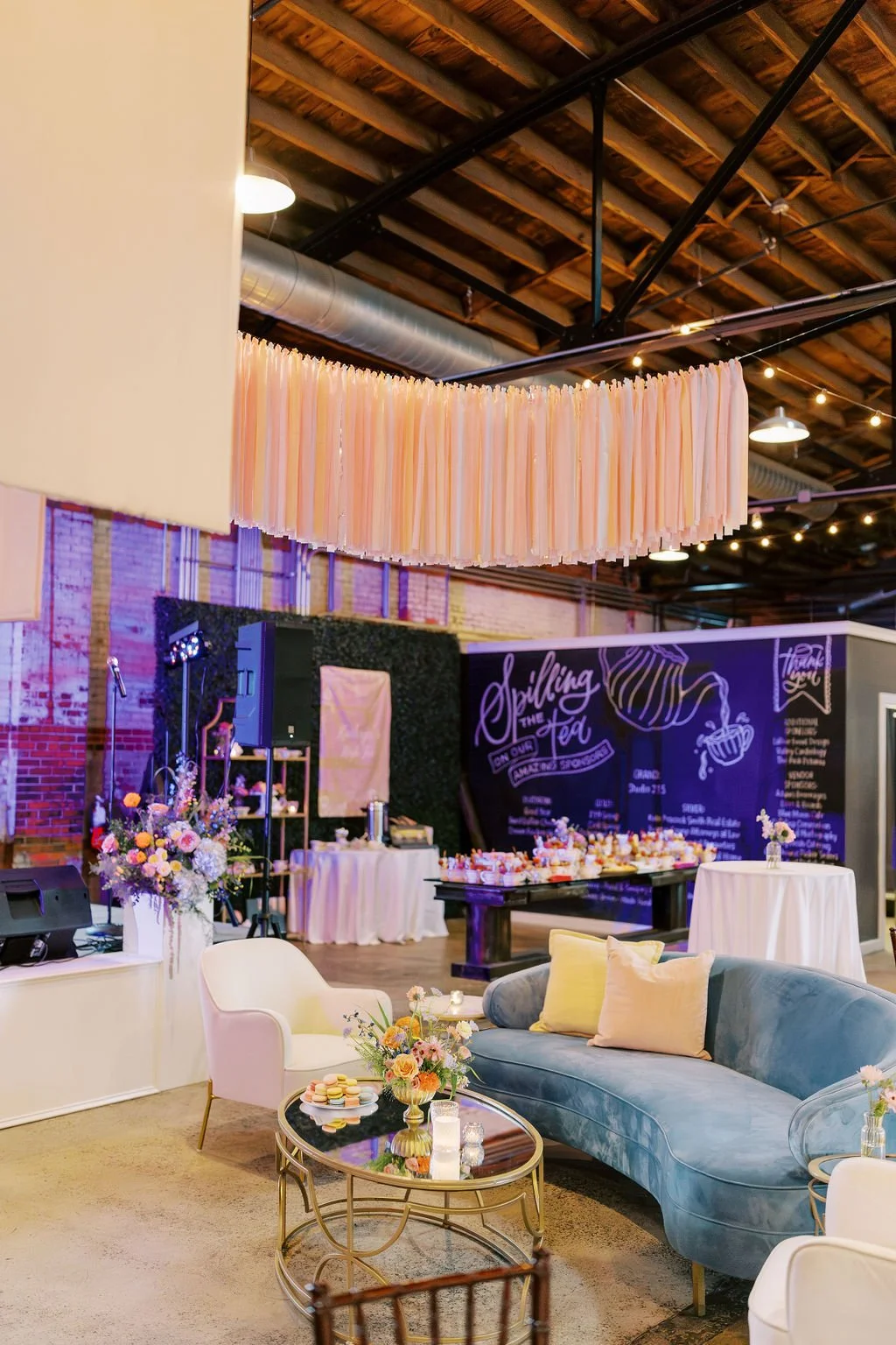 Interior of a decorated venue with a pink ribbon-like hanging decoration, floral arrangements, a velvet sofa, a white armchair, and a table with macarons, with a chalkboard wall in the background reading 'Spilling the Tea'.