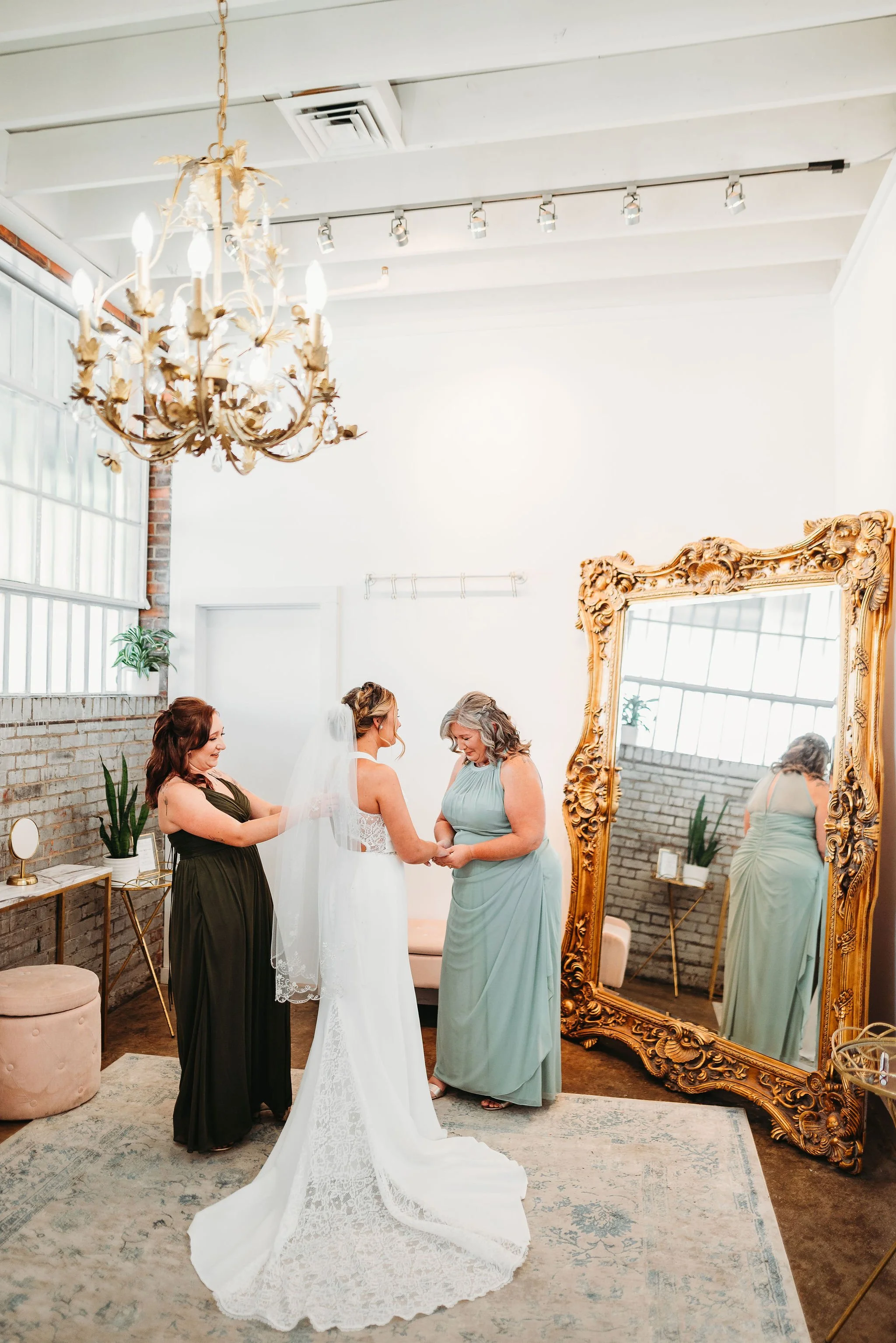 A bride in a white wedding dress and veil standing with two women, one helping with her dress. The setting is a bright, elegant room with a large ornate mirror, a chandelier, and indoor plants.