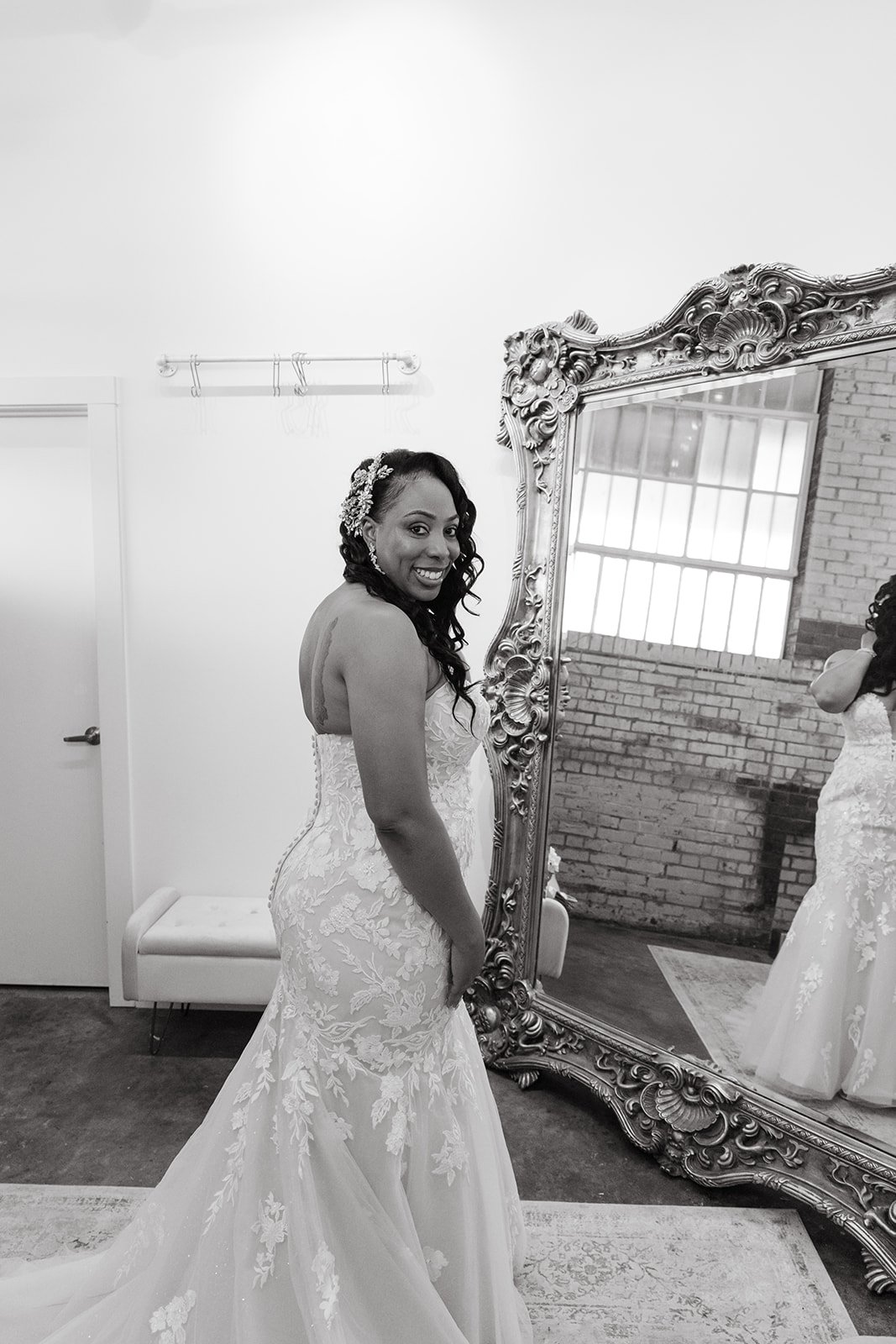 A woman in a wedding dress standing in front of a decorative mirror, smiling at her reflection.