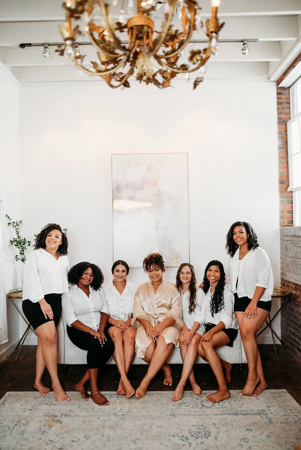 A group of seven women sitting and standing in a bright, modern room with white walls, a chandelier, and a large abstract painting. They are dressed in casual and semi-formal attire, smiling and posing for the photo.