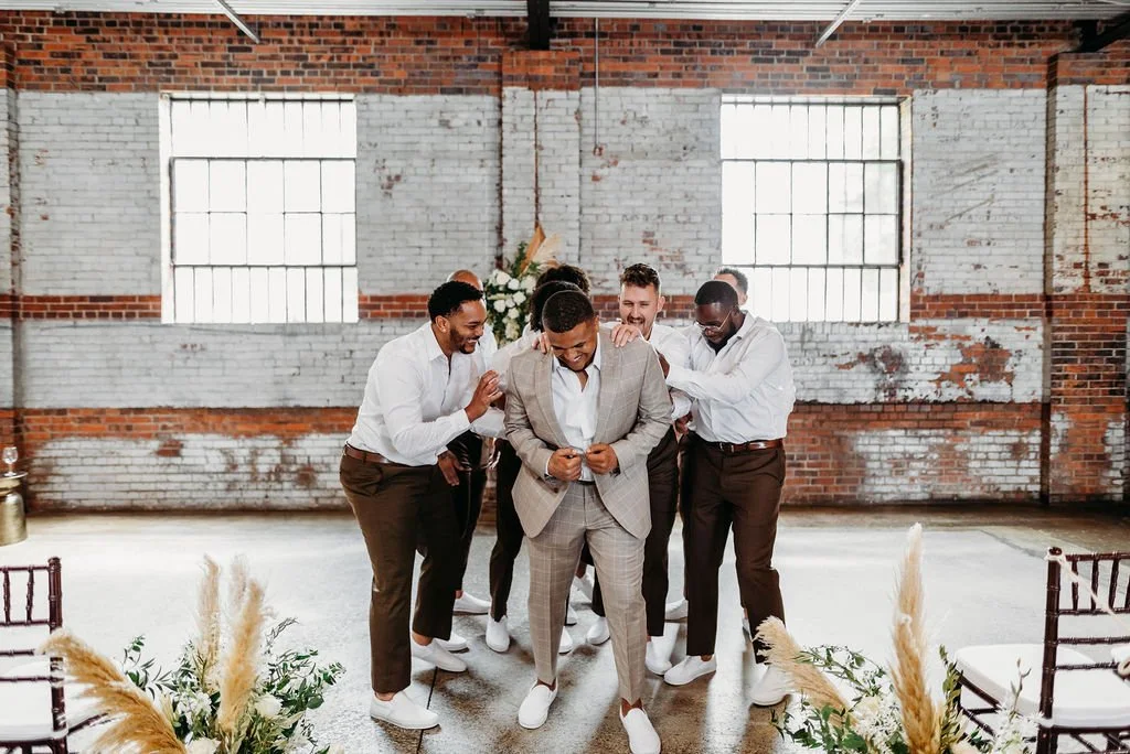 A group of men dressed in formal attire celebrating indoors with brick walls and large windows, surrounded by floral arrangements.