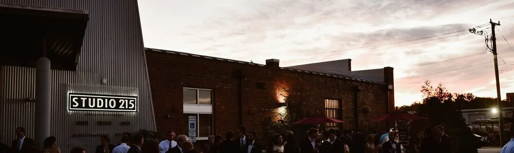 People gathered outside a brick building with a sign that reads 'STUDIO 215' during sunset.
