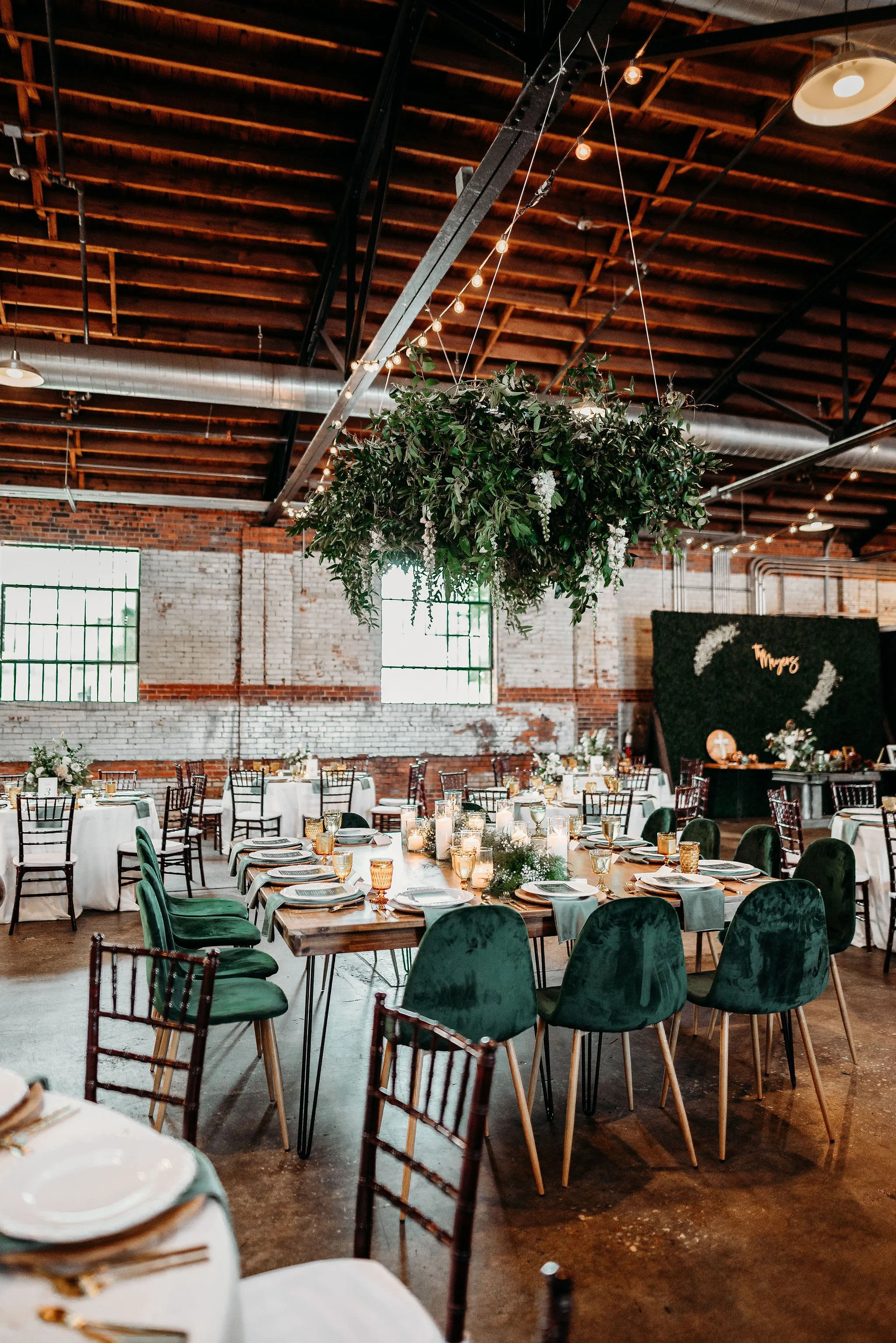 Indoor wedding reception with rustic industrial decor featuring round and rectangular tables set with white tablecloths, candles, floral centerpieces, and dark green velvet chairs, with a large hanging floral arrangement above.