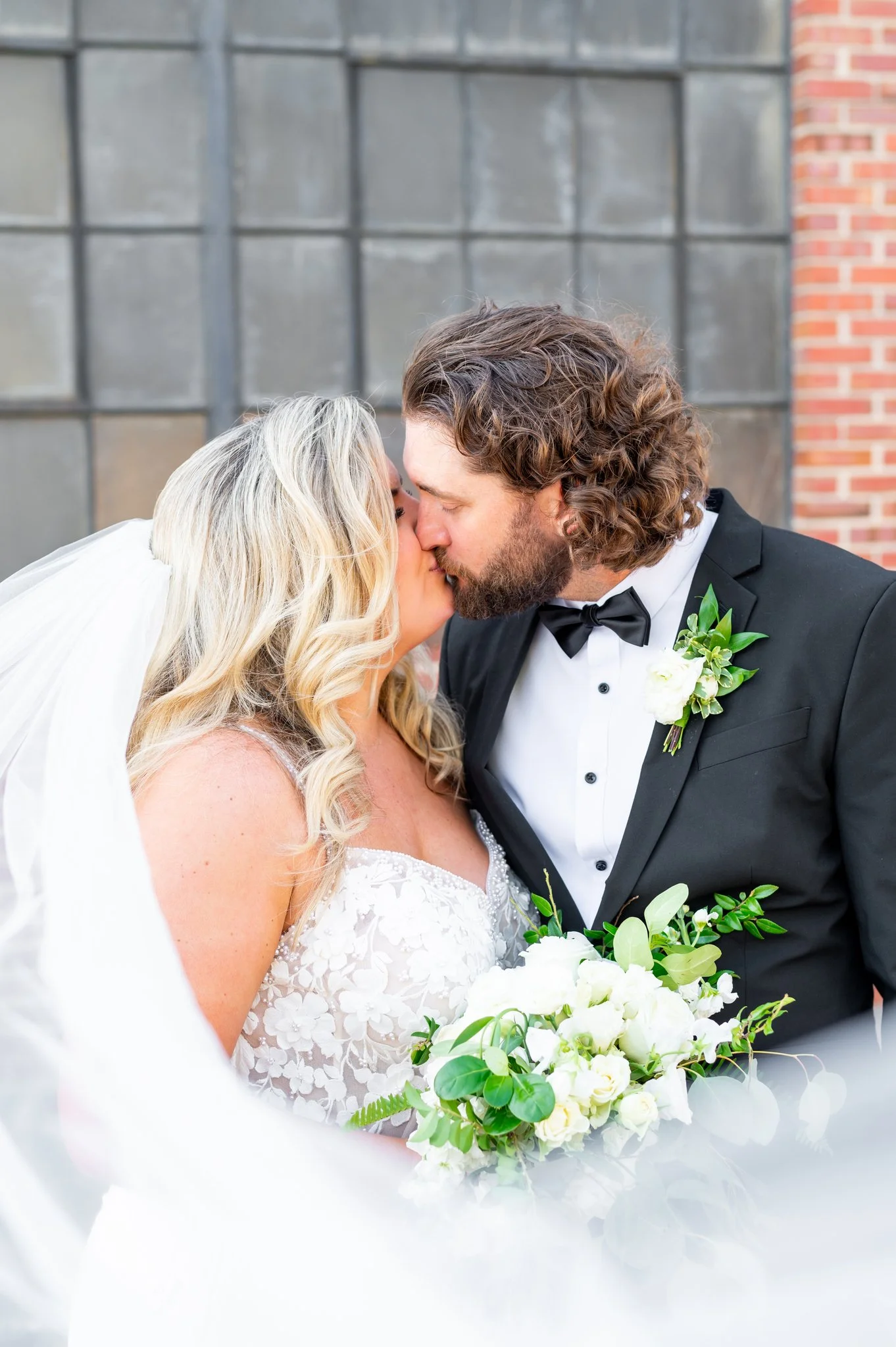 A bride and groom sharing a kiss on their wedding day, with the bride holding a bouquet of white flowers and the groom in a tuxedo, standing outdoors in front of a brick and metal wall.