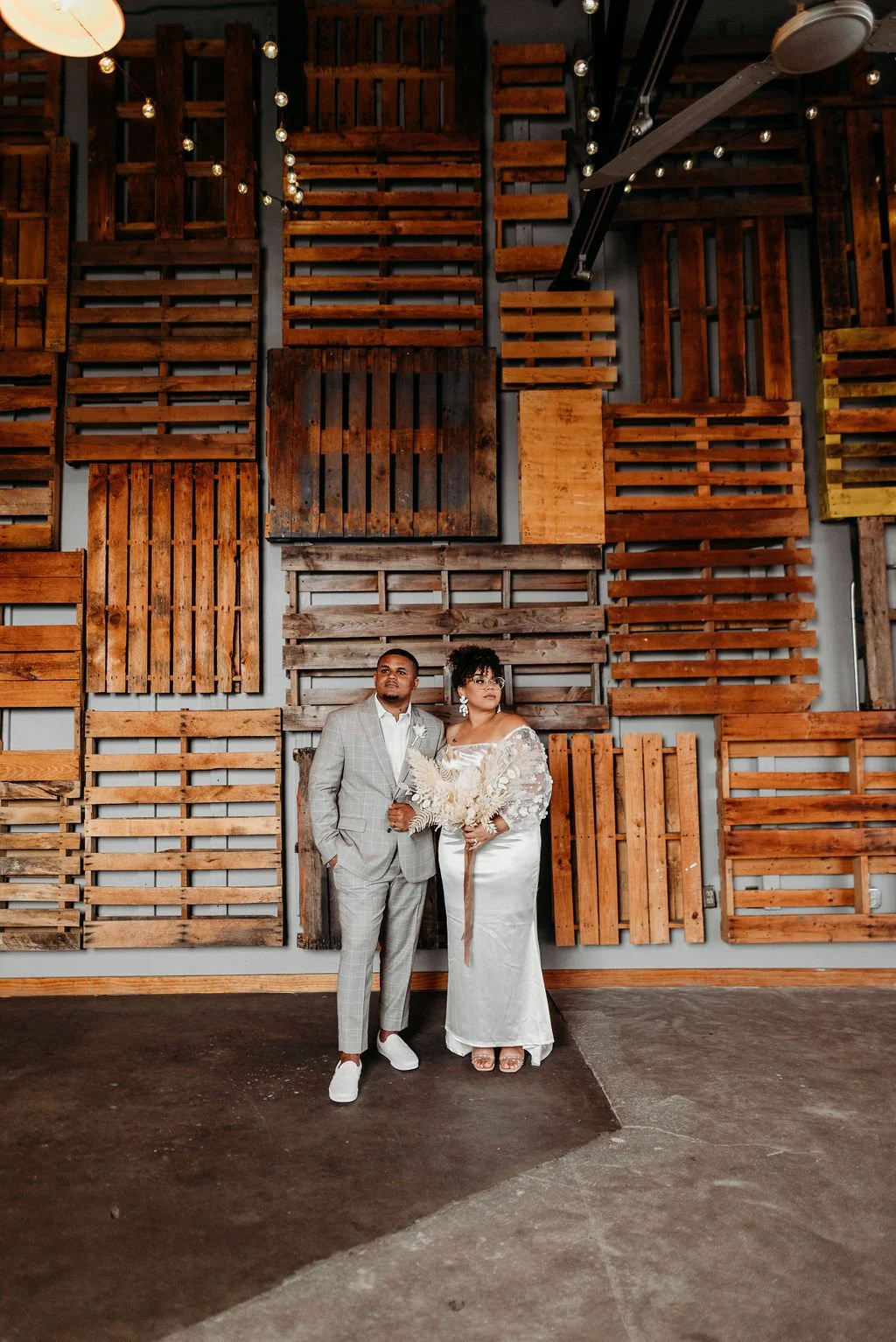 A couple standing in front of a wall decorated with wooden pallets, holding hands, dressed in wedding attire.