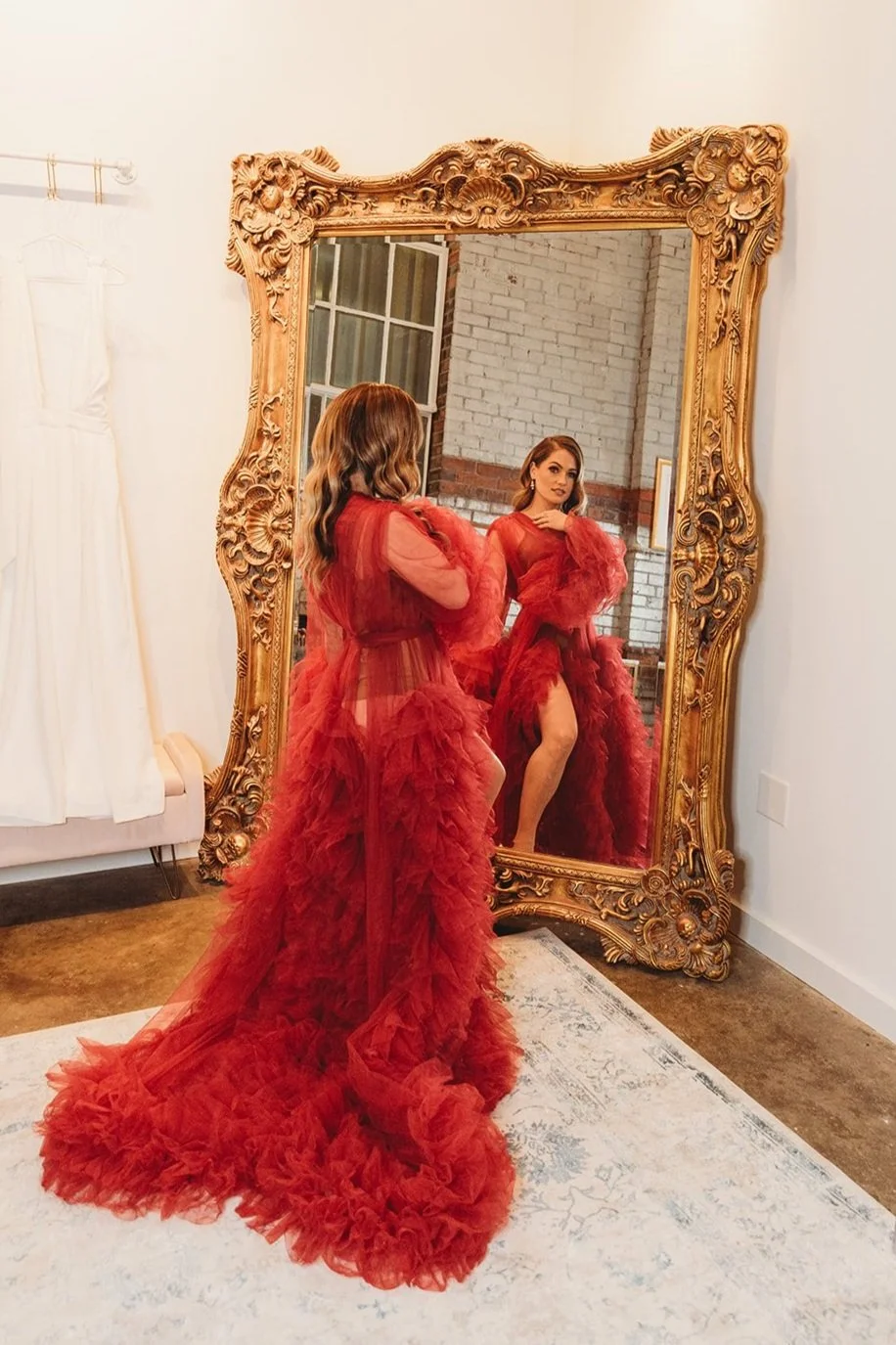 Woman in a red, ruffled, sheer dress looking at her reflection in an ornate gold mirror in a room with brick wall and large window.