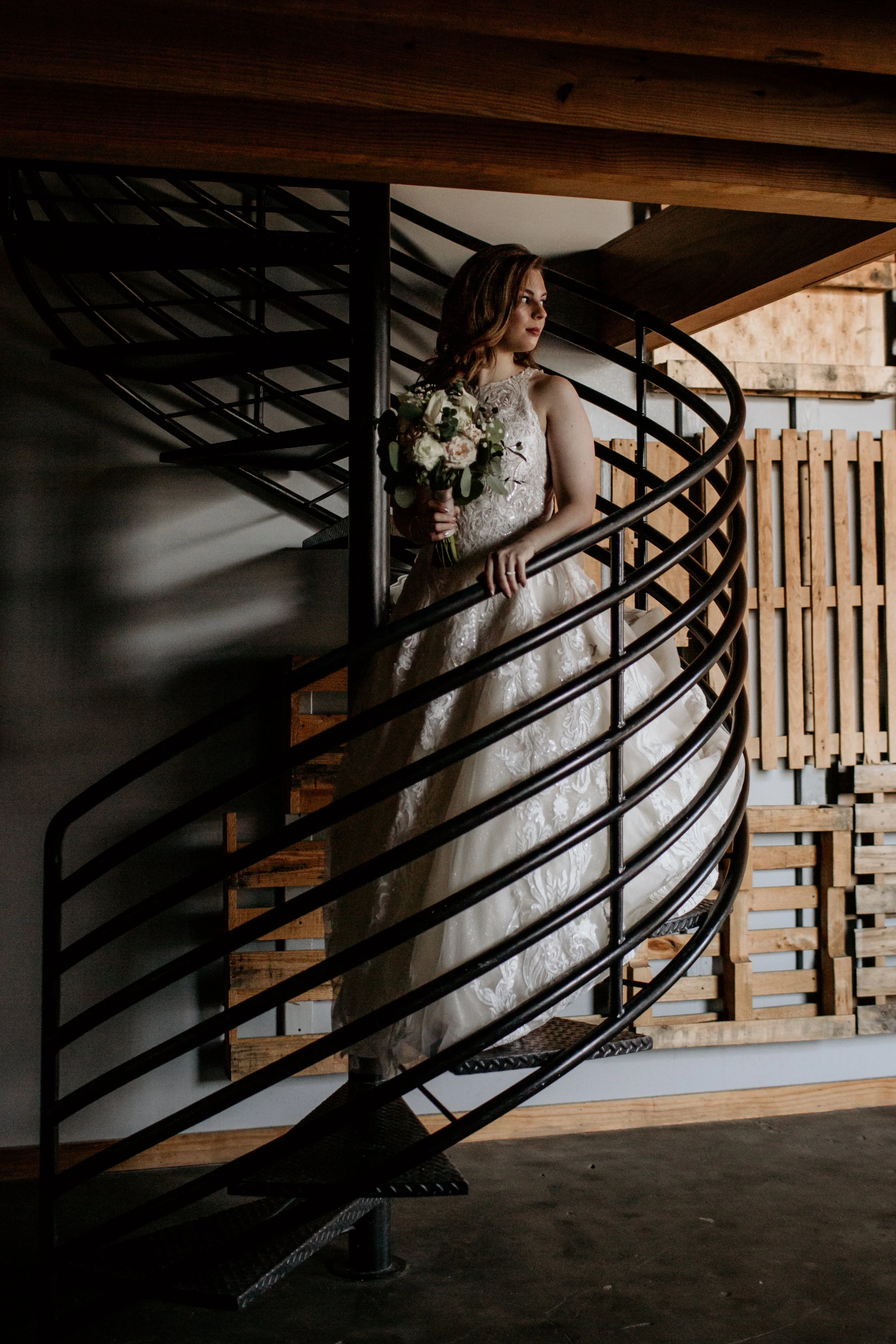 A woman in a wedding dress standing on a spiral staircase, holding a bouquet of flowers, looking to her side in an indoor setting with wood and metal decor.