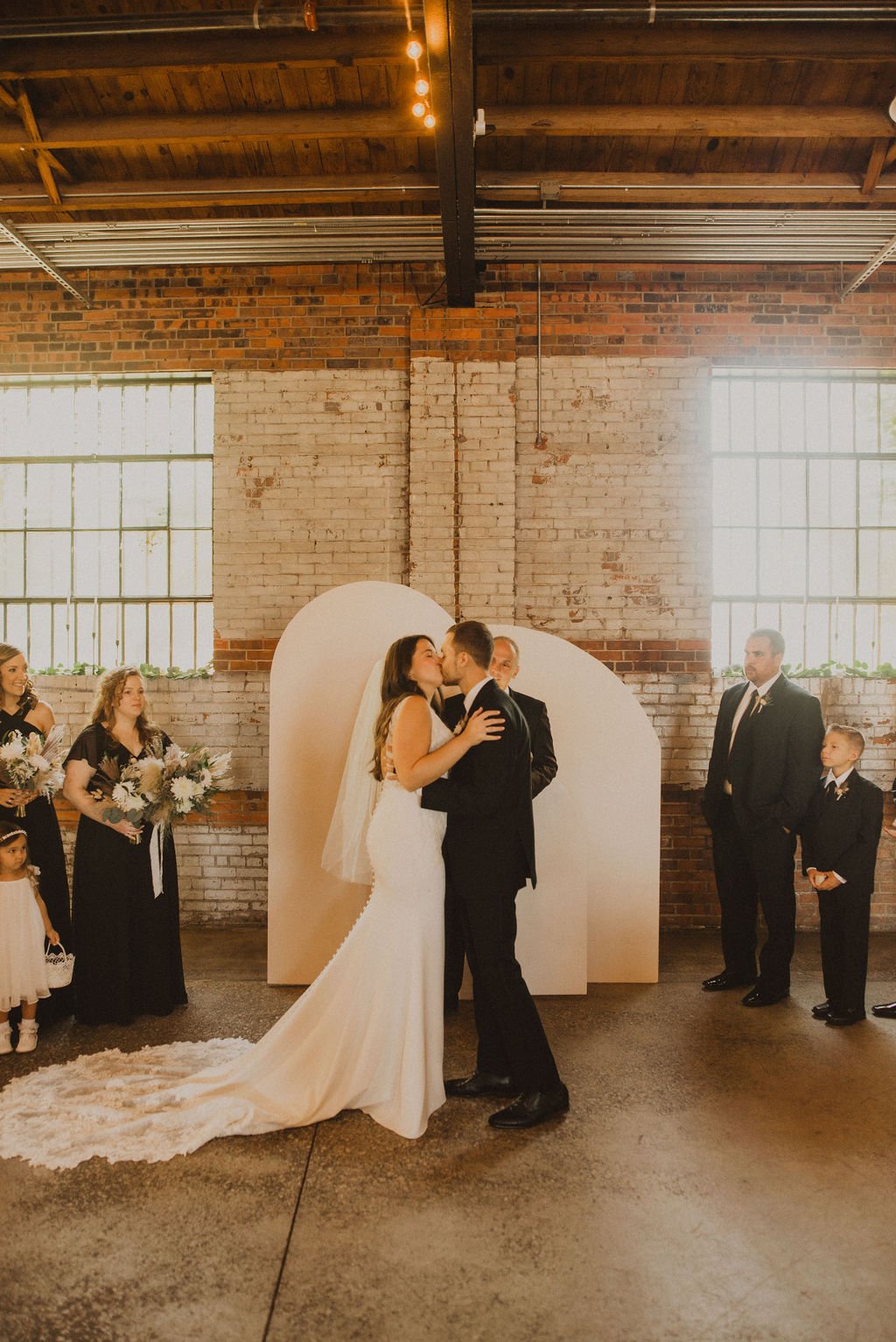 A wedding ceremony with a bride and groom kissing, surrounded by wedding party members in a rustic venue with brick walls and large windows.
