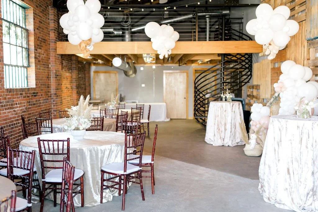 Elegant event space decorated with white balloons and floral arrangements, with round tables covered in white tablecloths and dark wood chairs, exposed brick walls, large windows, and a spiral staircase.