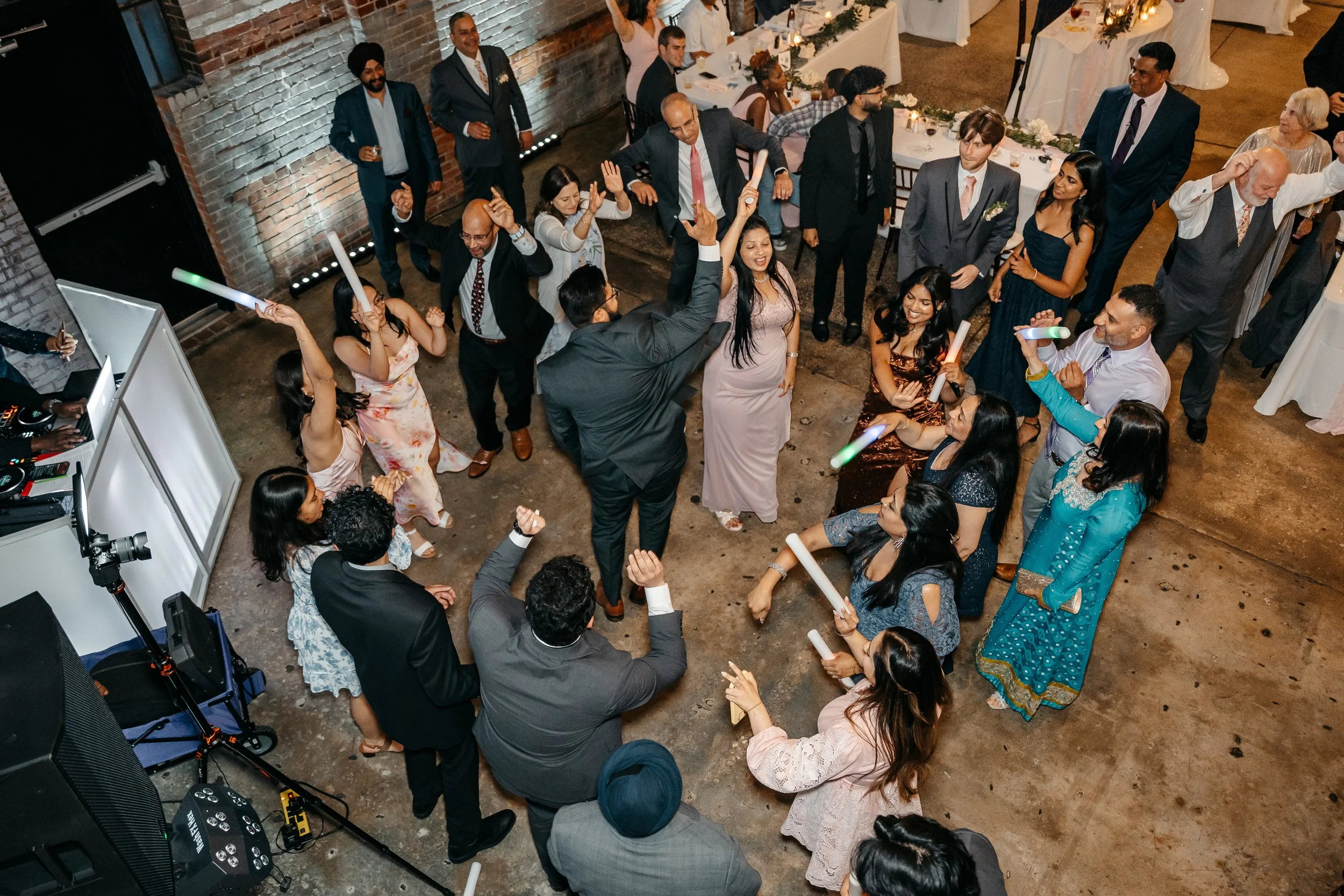 Guests dancing and celebrating at a wedding reception with fairy lights, tables, and a DJ setup in an industrial-style venue.