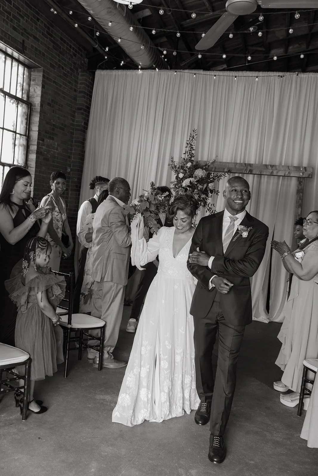 A bride and groom walking down the aisle of their wedding ceremony, smiling and holding hands, with guests clapping and taking pictures around them.