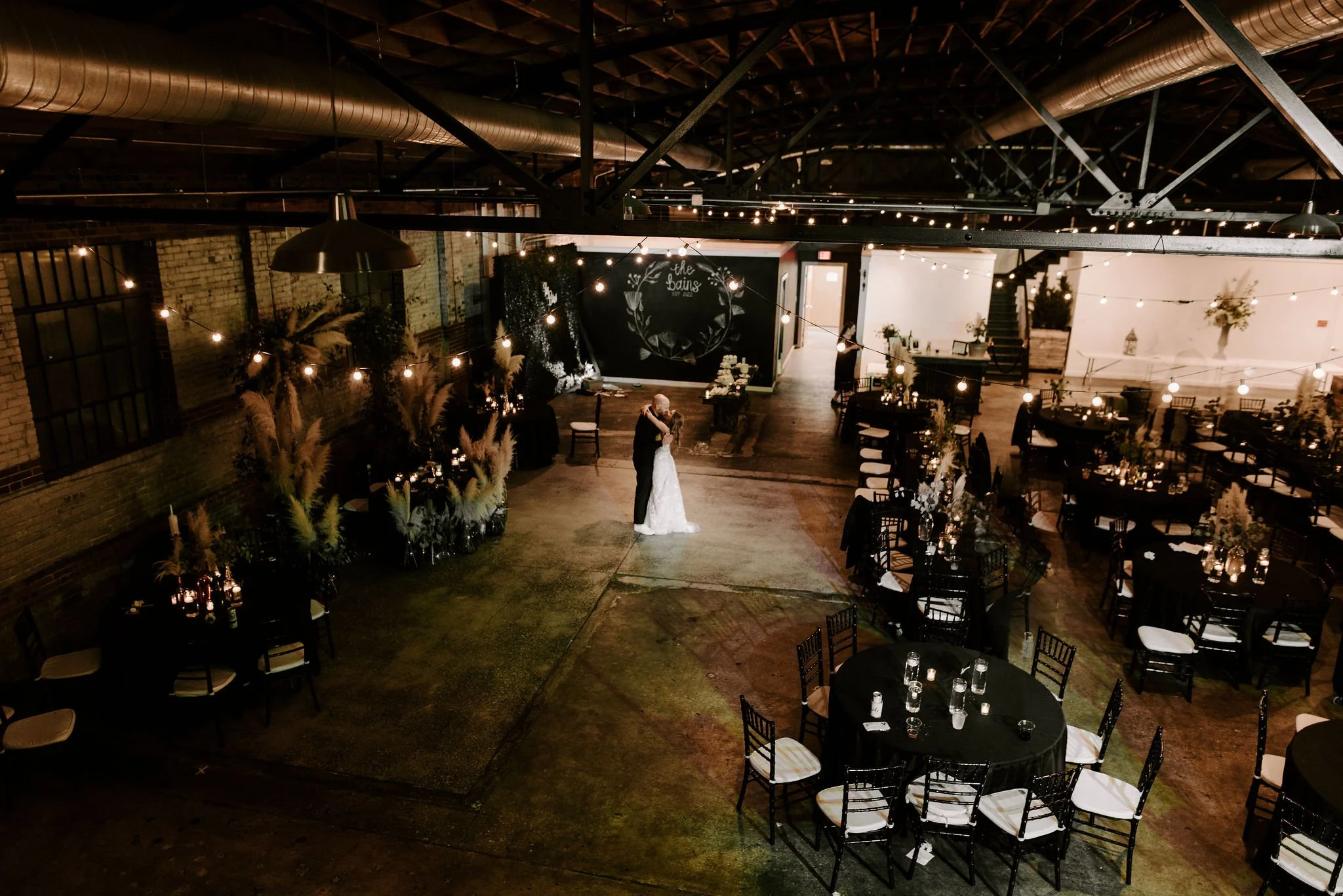 A bride and groom dancing in a decorated event space with round tables, black tablecloths, floral centerpieces, and fairy lights hanging from the ceiling.