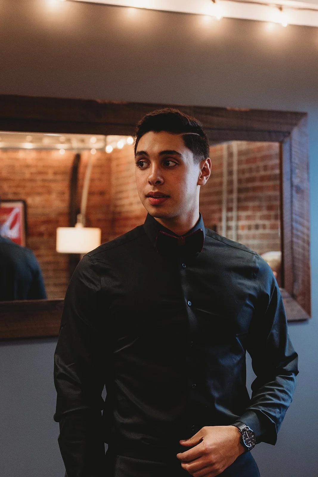 Young man in black dress shirt and bow tie standing indoors against a brick wall with a mirror, warm lighting, and a lamp reflected in the mirror.