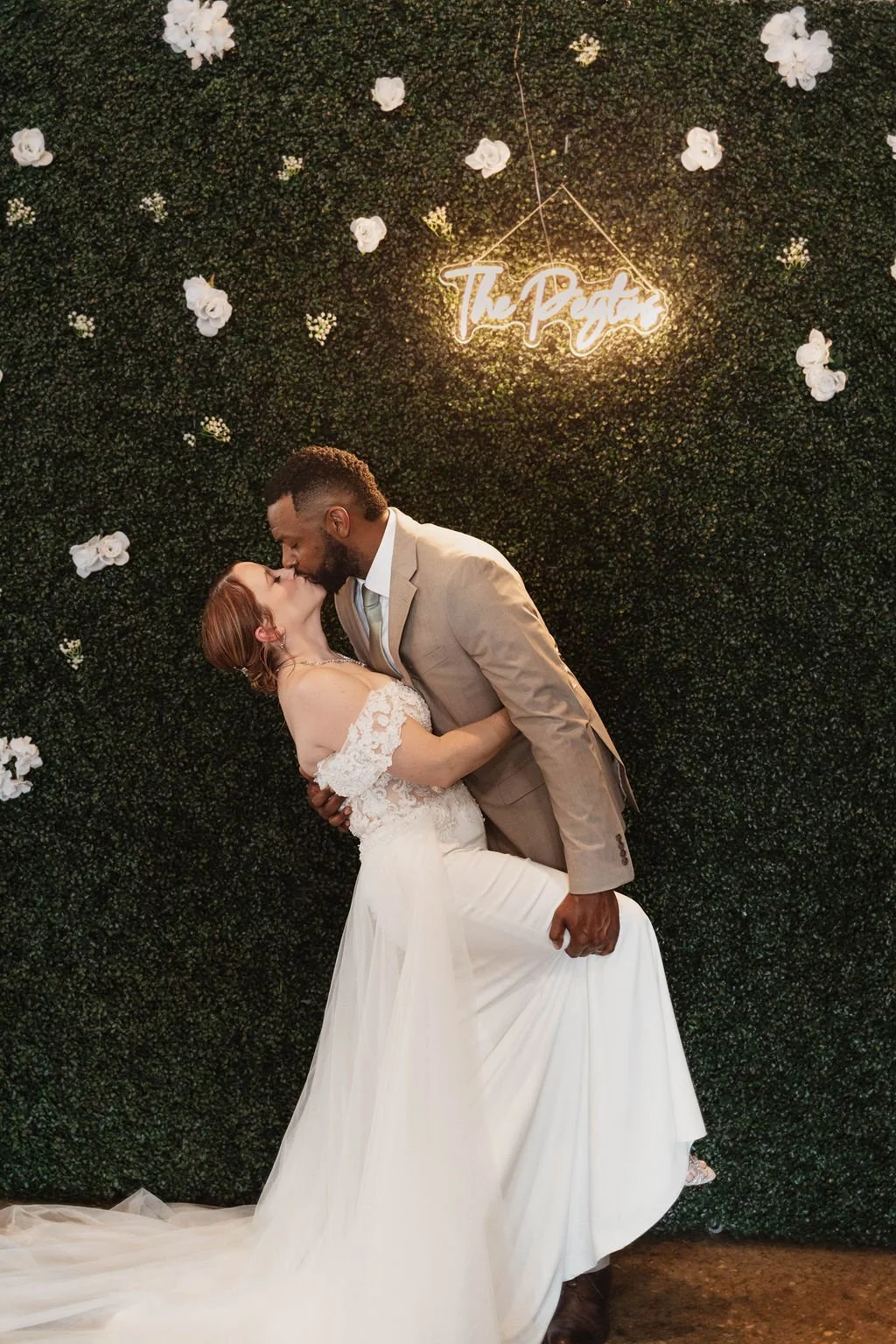 A couple kissing at their wedding, with a decorative green wall background, white flowers, and a neon sign that says 'The Party'.