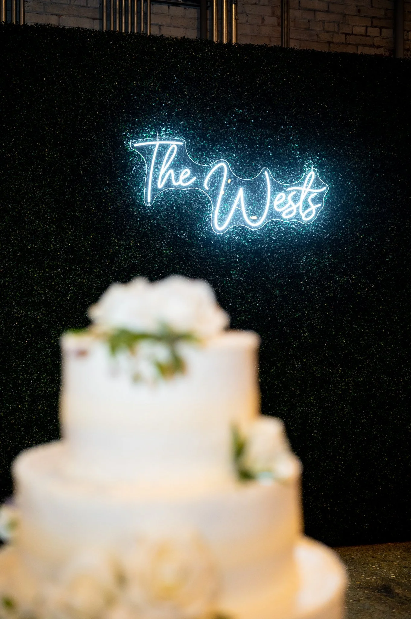 Neon sign reading 'The WEst' in cursive font, mounted on a black glittery backdrop, with a blurred white wedding cake with flowers in the foreground.