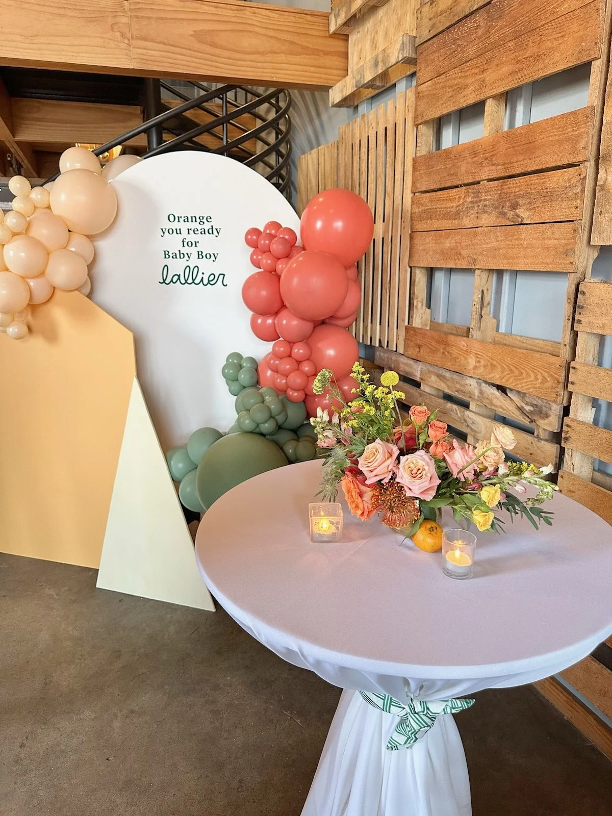 Decorative balloon arrangement with pink, green, peach, and beige balloons near a sign that reads, "Orange you ready for Baby Boy Lallier," alongside a table with a flower bouquet and candles for a baby shower.