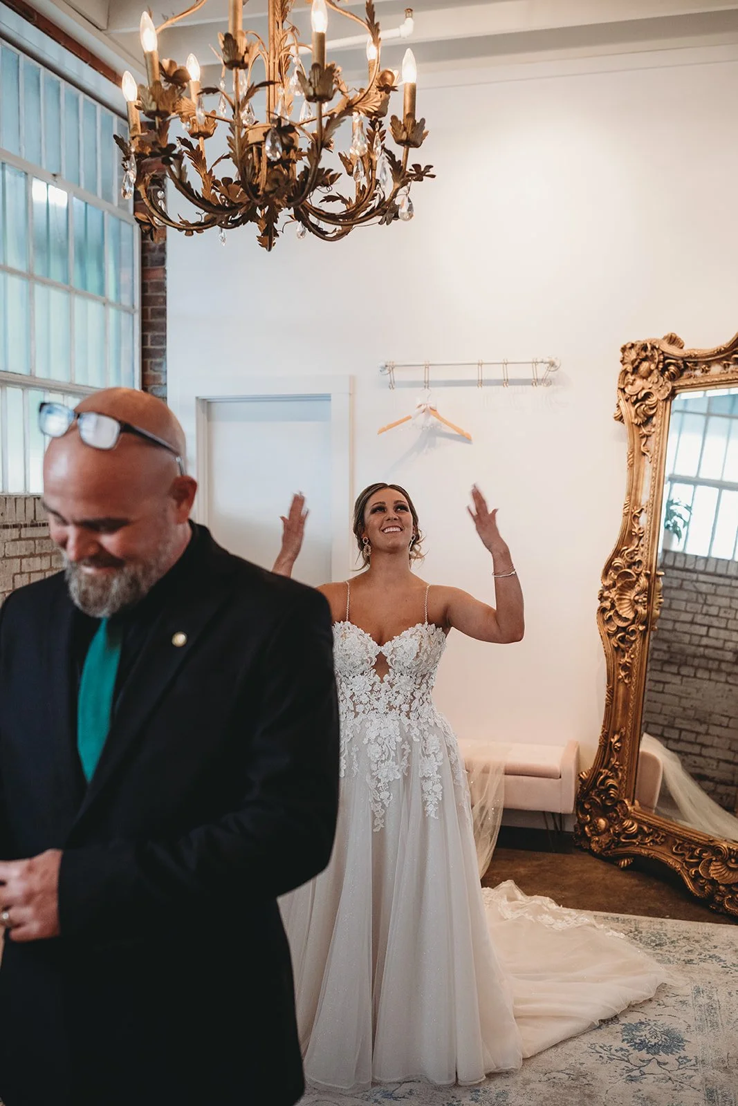 A bride in a lace wedding dress stands with her arms raised and a joyful expression, in a room with a chandelier and a large ornate mirror, while a man in a black suit with sunglasses on his head smiles in the foreground.