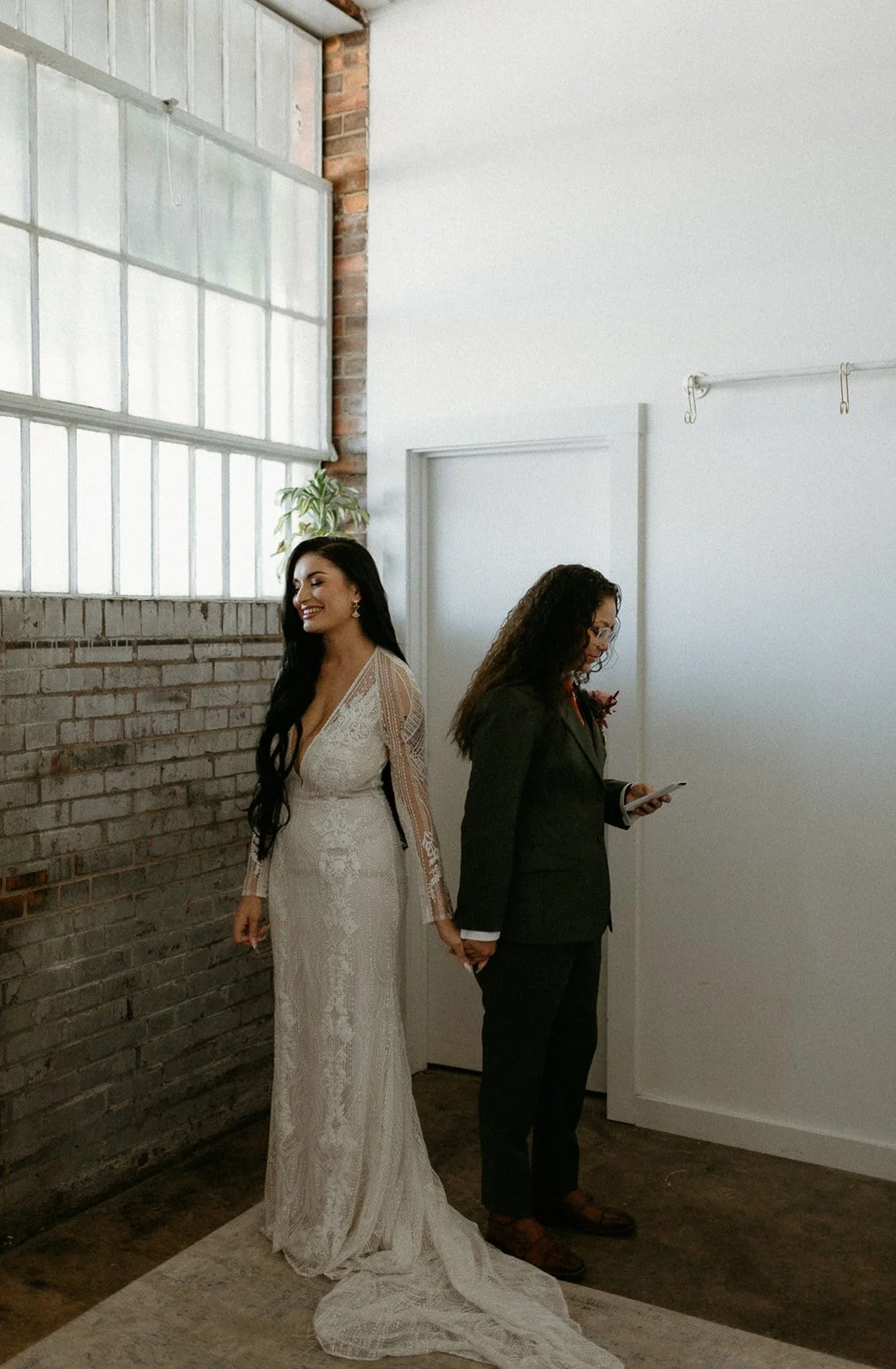 A bride and her partner holding hands during a wedding ceremony indoors. The bride is wearing a long, lace wedding dress and has long dark hair, while the woman is dressed in a dark suit and is looking at her phone.