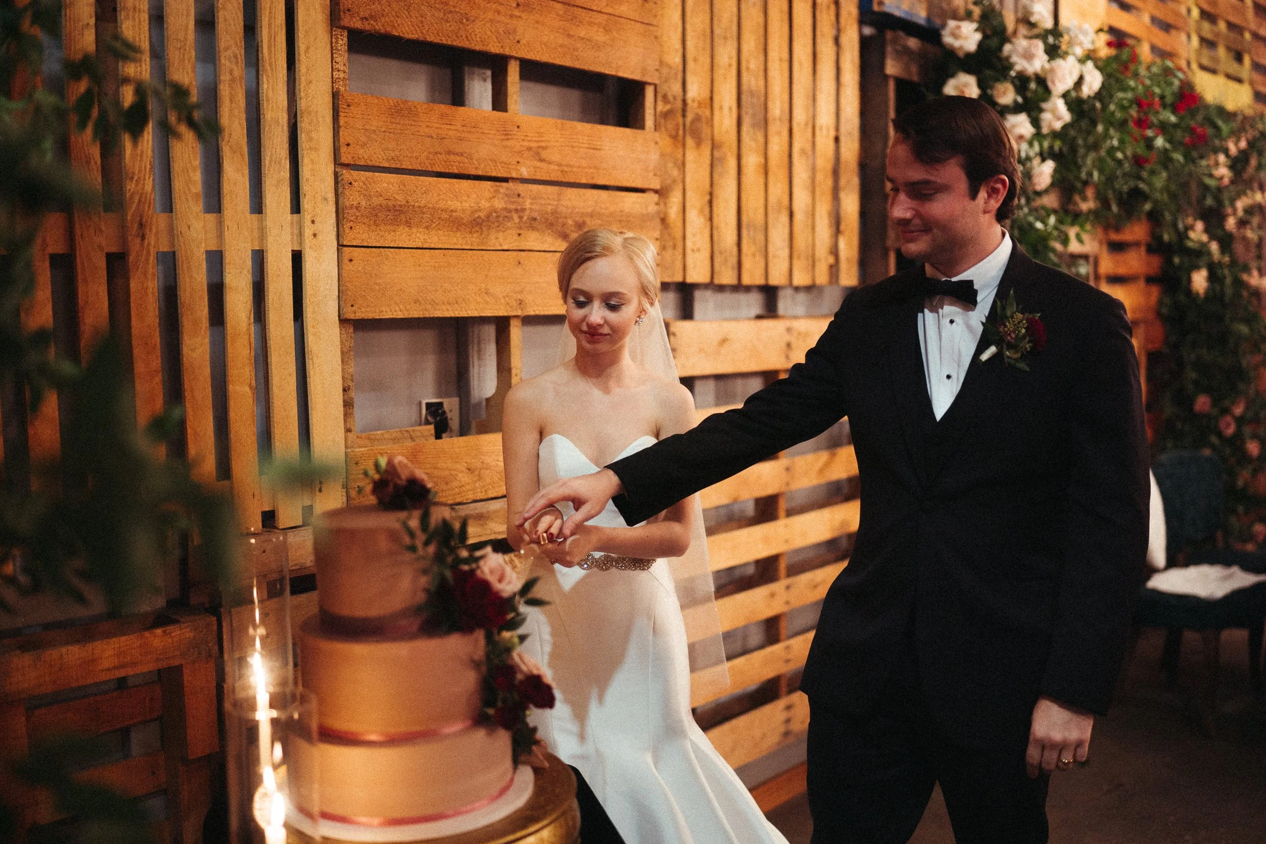 A bride and groom cutting a wedding cake indoors. The bride is wearing a white strapless wedding gown and veil, and the groom is in a black tuxedo with a bow tie. The cake is decorated with flowers.