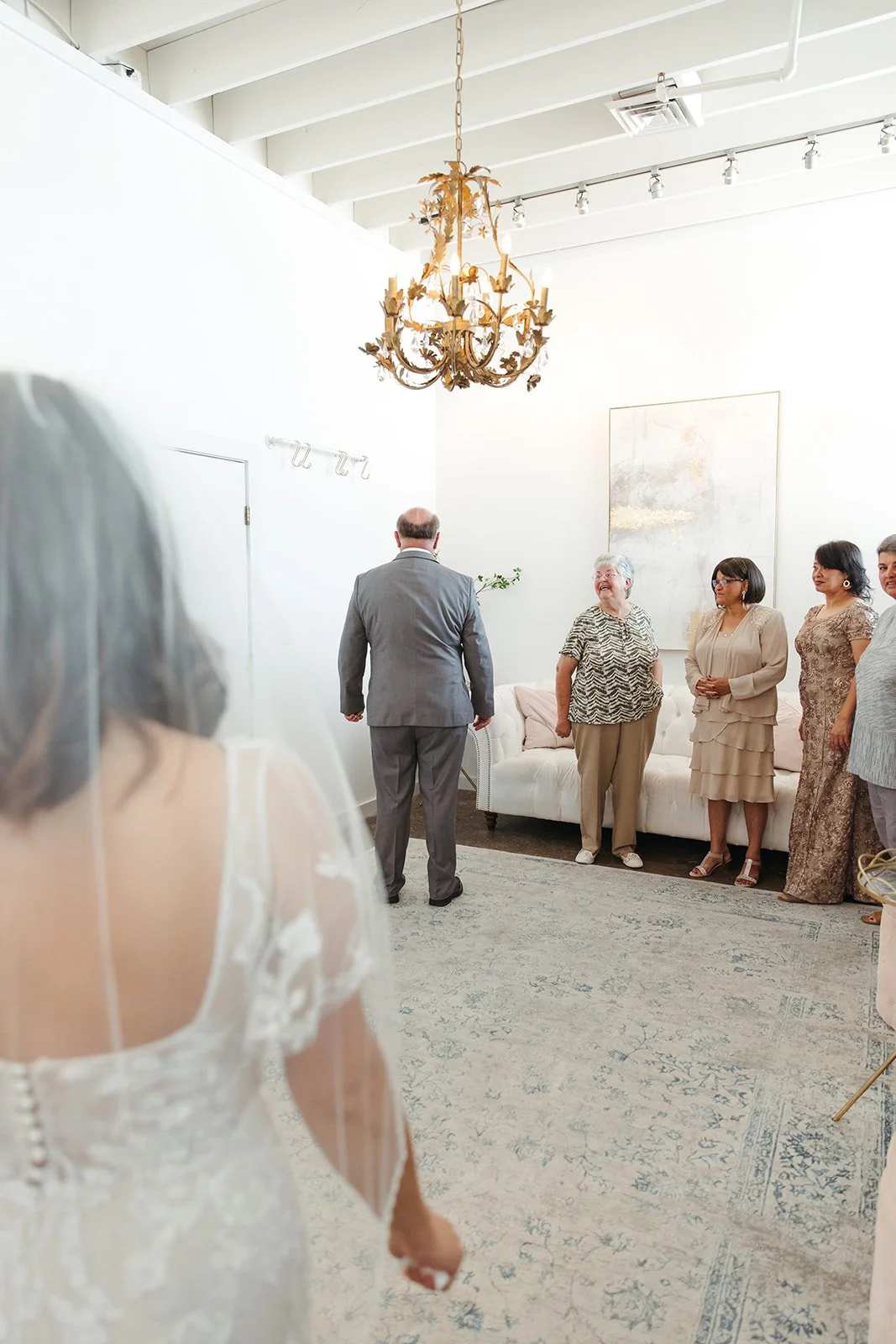 A wedding ceremony with a bride in a white dress with lace details standing on the left, facing a group of guests in a bright, minimalist room. The guests, mostly women, are standing and smiling, with a man in a gray suit at the center. The room has white walls, a large abstract painting, a vintage chandelier, and a plush white sofa.