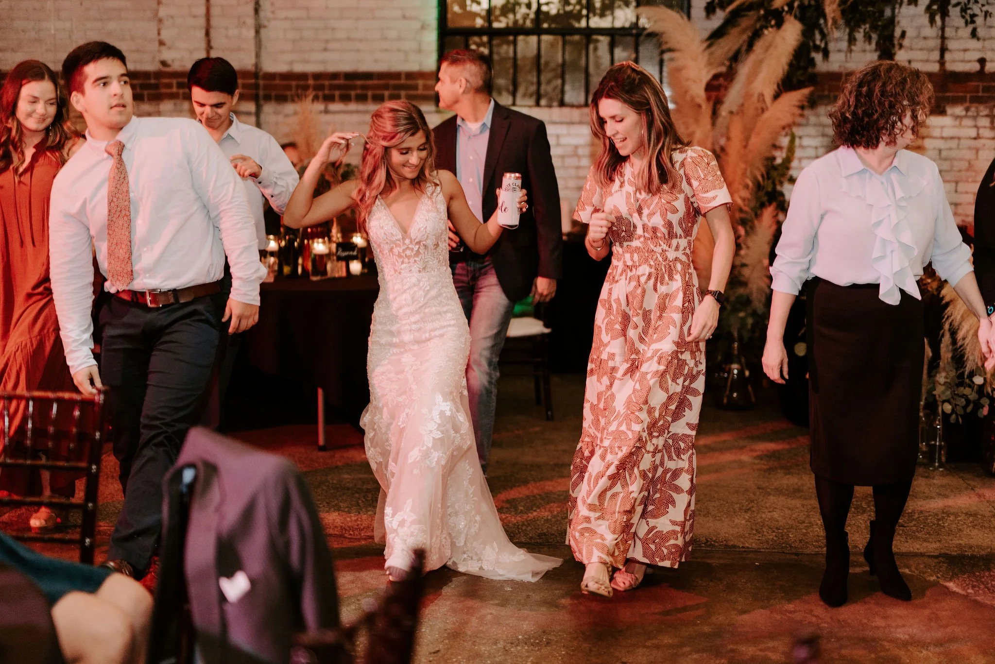 Group of people dancing at a wedding reception, with a bride in a white lace wedding dress and several women in dresses, in a rustic venue with brick walls and tall pampas grass decorations