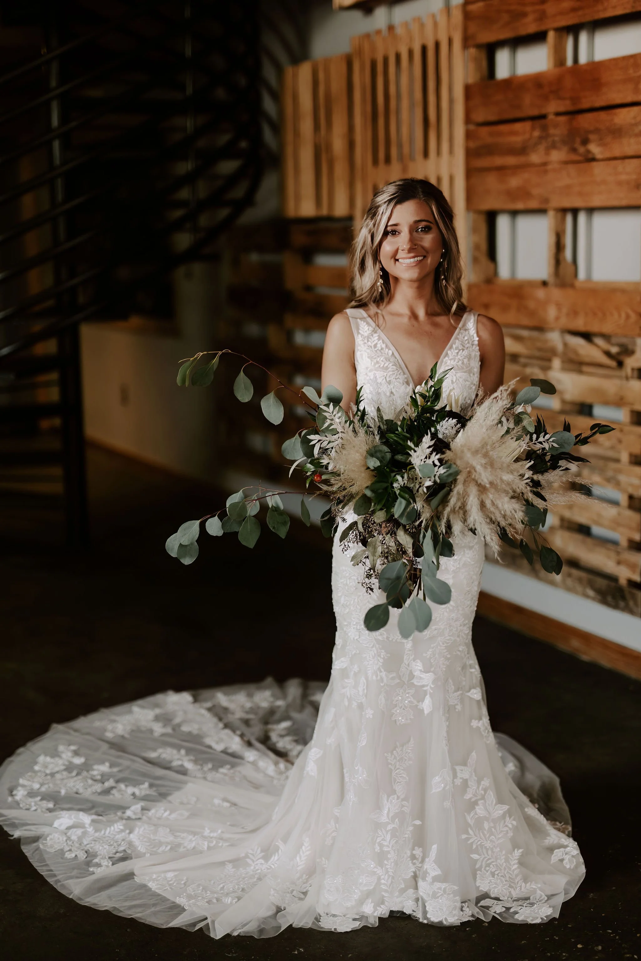 A bride in a white lace wedding gown holding a large bouquet of greenery and pampas grass, standing indoors with wooden wall decor.