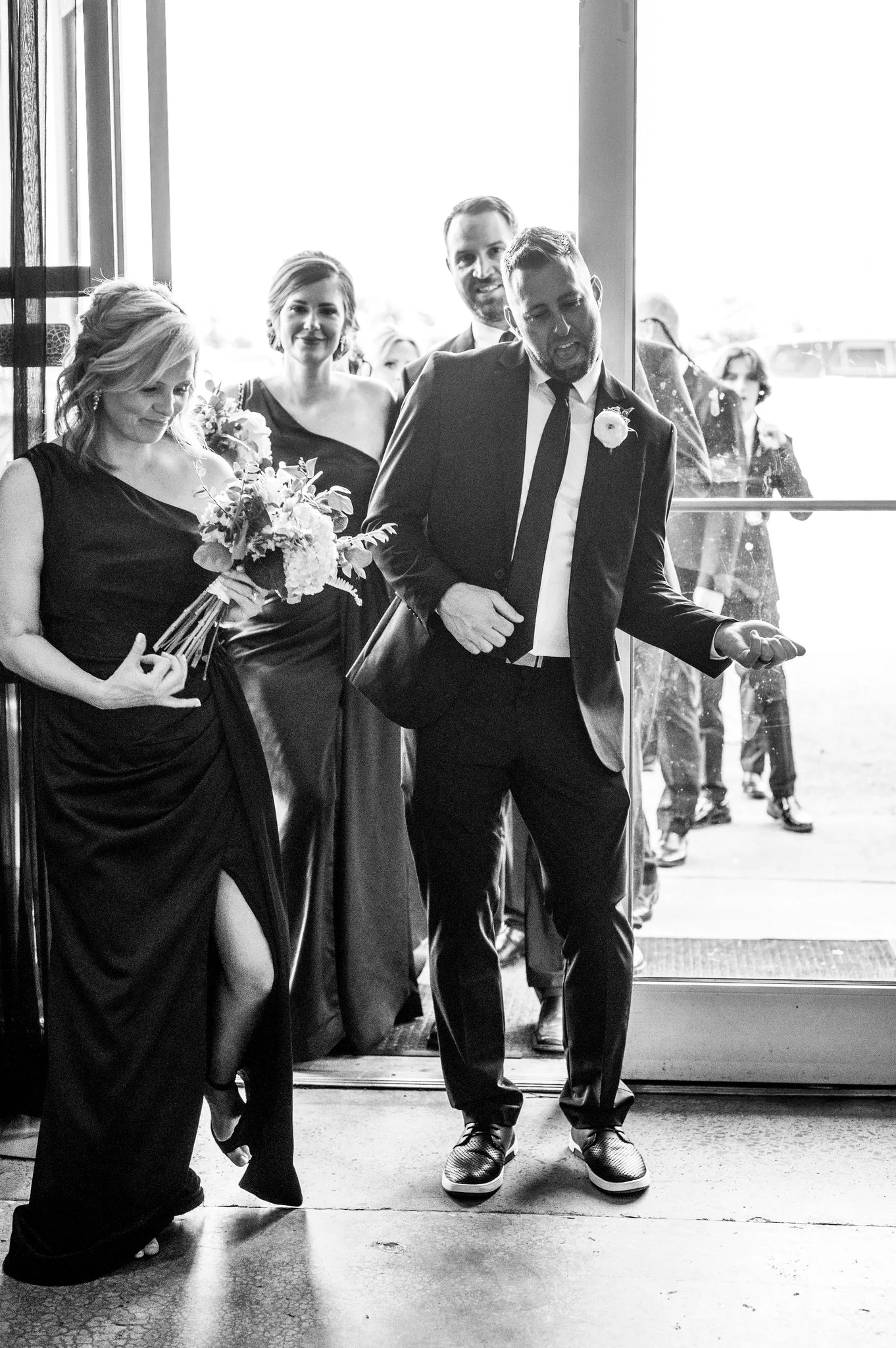 Black and white photo of a wedding party exiting a building, including a woman holding a bouquet, a man in a suit celebrating, and others behind them.