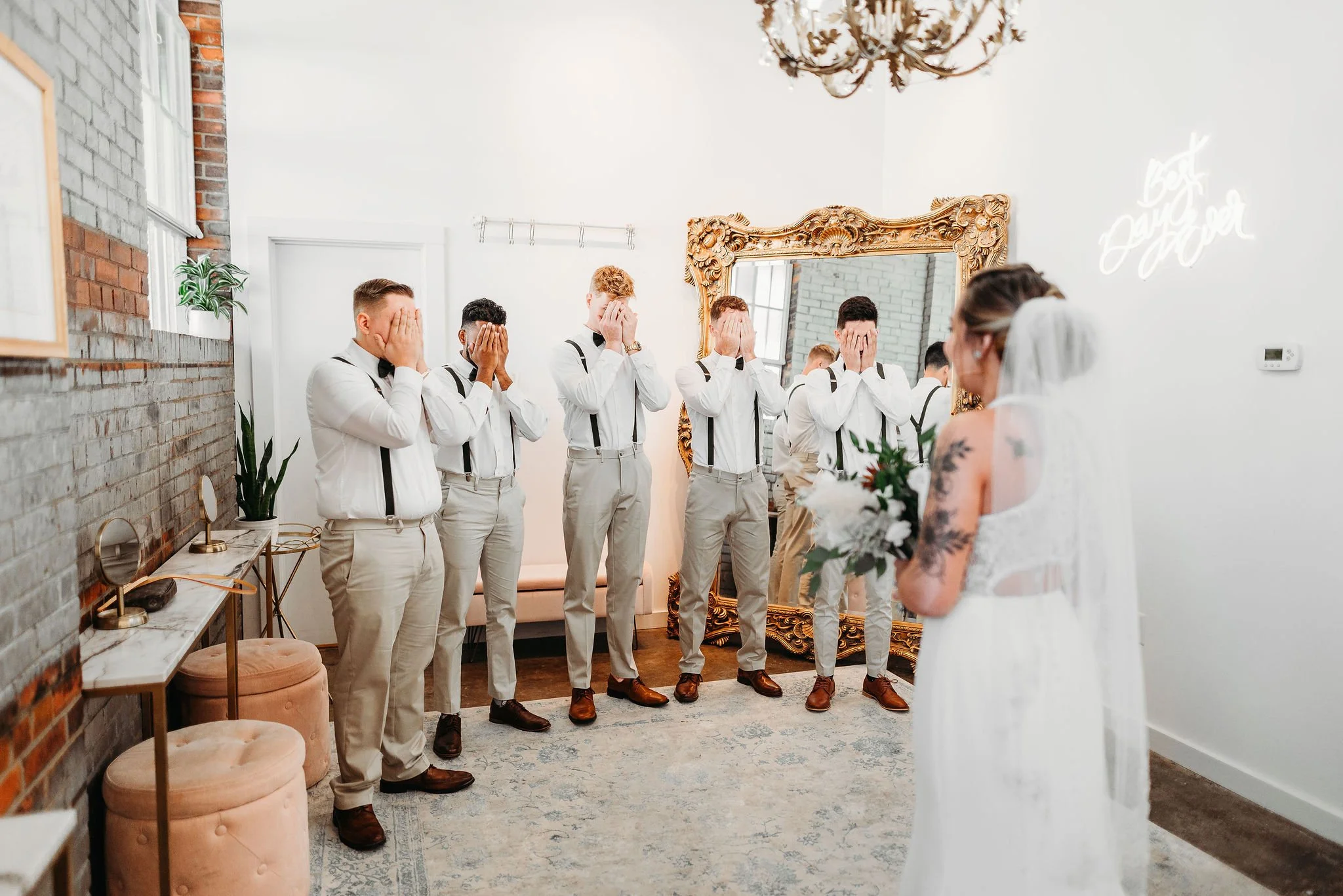 Bride in white wedding dress with veil holding bouquet, facing six groomsmen with hands covering faces, in a room with white walls, large ornate mirror, neon sign, and brick accent wall.