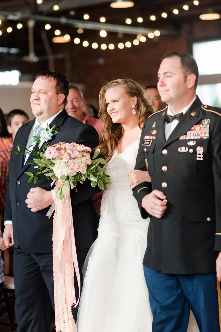 A bride in a white wedding dress holding a bouquet of pink and white flowers, walking down the aisle with a man in a dark suit, two men in military uniforms on either side, and guests watching in the background. The setting has warm lighting and string lights overhead.