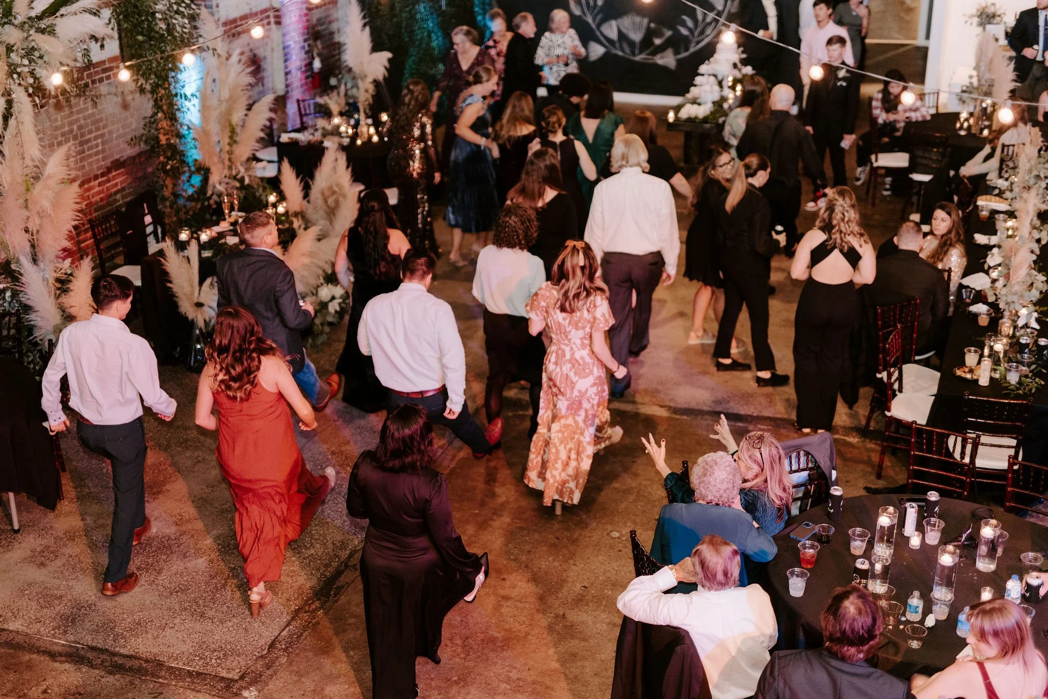 People dancing and socializing at a celebration or party in a decorated indoor venue with tables, candles, and string lights.