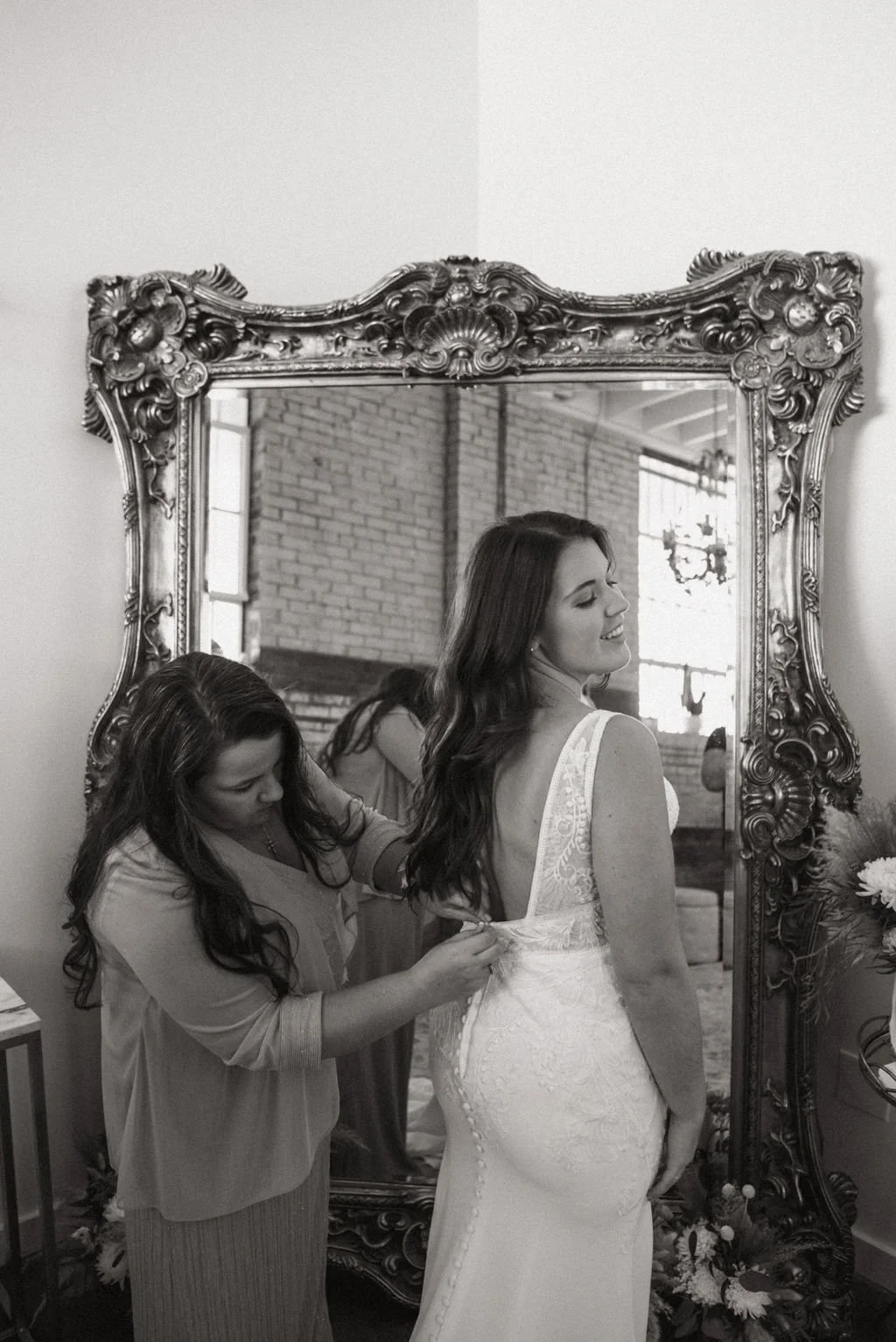 A woman in a wedding dress smiling in front of a large ornate mirror while another woman helps adjust her dress.