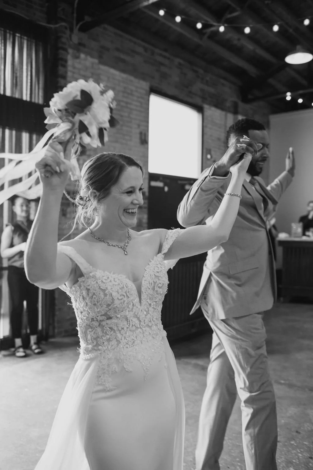 A bride and groom celebrating, holding hands up with joyful expressions in an indoor wedding venue.