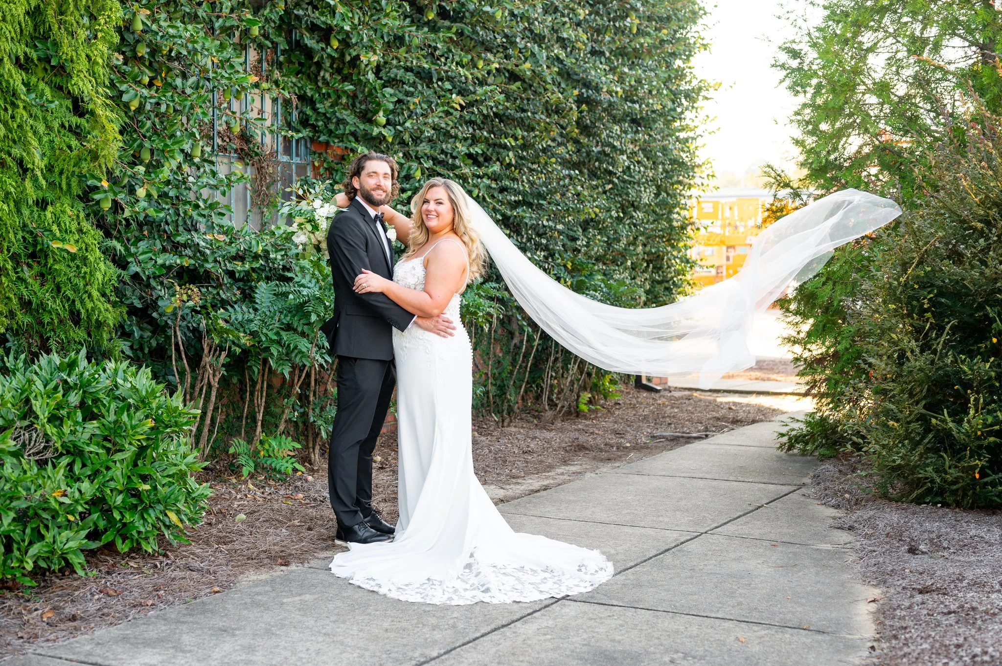 A newlywed couple stands together on a sidewalk surrounded by lush greenery. The bride wears a white lace wedding gown with a long veil flowing in the breeze. The groom is dressed in a black tuxedo. They are smiling and embracing, celebrating their w