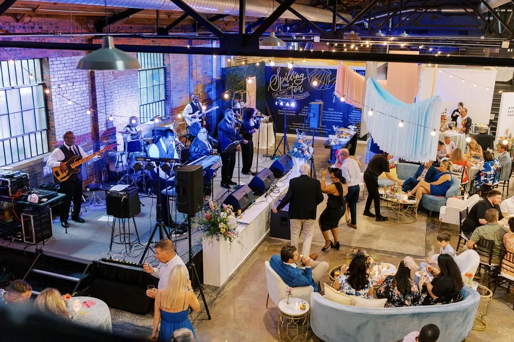 People dancing and socializing at a wedding reception with a live band on stage and decorated with string lights and floral arrangements.
