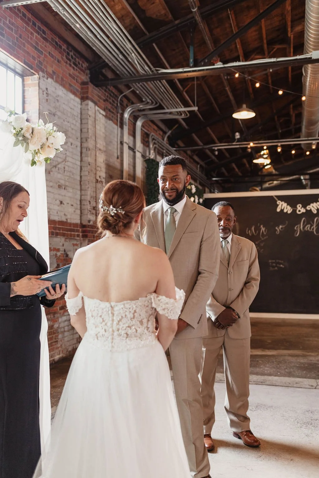 A couple gets married indoors in a rustic venue with brick walls and wooden ceiling beams. The bride wears an off-shoulder lace wedding dress with her hair in an updo, and the groom wears a tan suit with a light green tie. An officiant stands to the 