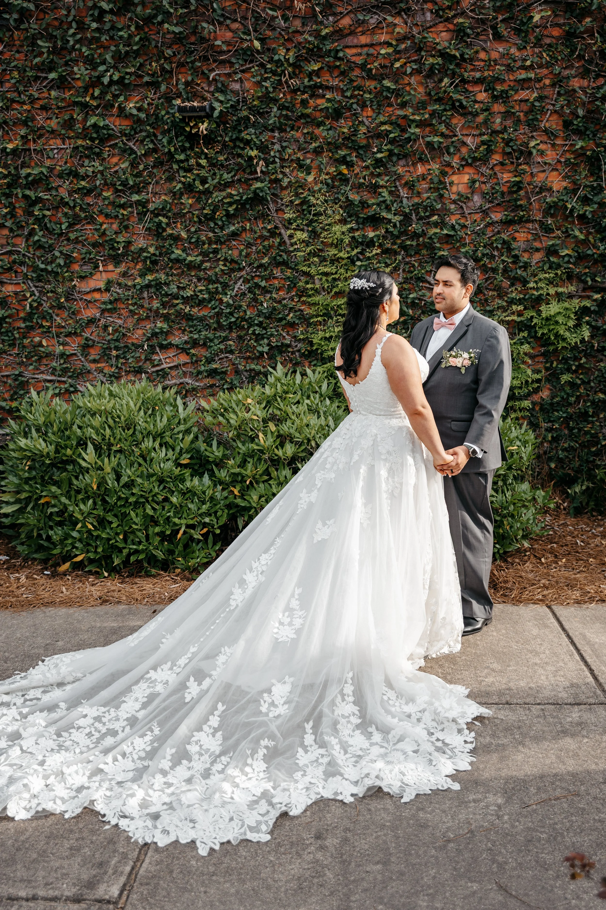 A bride in a white wedding gown with floral lace detailing holding hands with a groom in a gray suit with a pink bow tie, standing outdoors on a sidewalk with green bushes and a brick wall covered in ivy in the background.