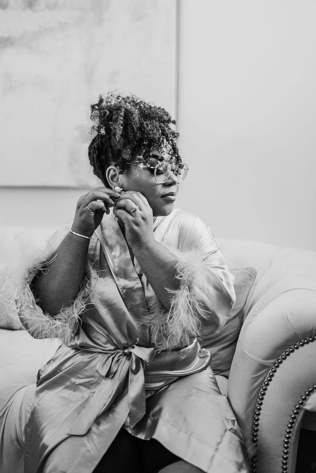 A woman with curly hair, glasses, and earrings is sitting on a sofa, adjusting her earring. She is wearing a satin robe with feathered cuffs, and appears to be in a dressing room or living room setting.