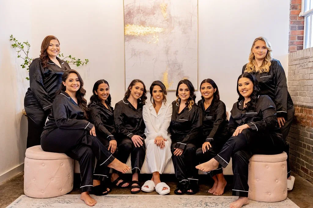 A group of nine women, dressed in matching black pajamas with white piping, sitting and standing on and around two pink ottomans in a modern room with a beige wall, an abstract painting, a brick wall, and a window. The woman in the center wears white pajamas with slippers.