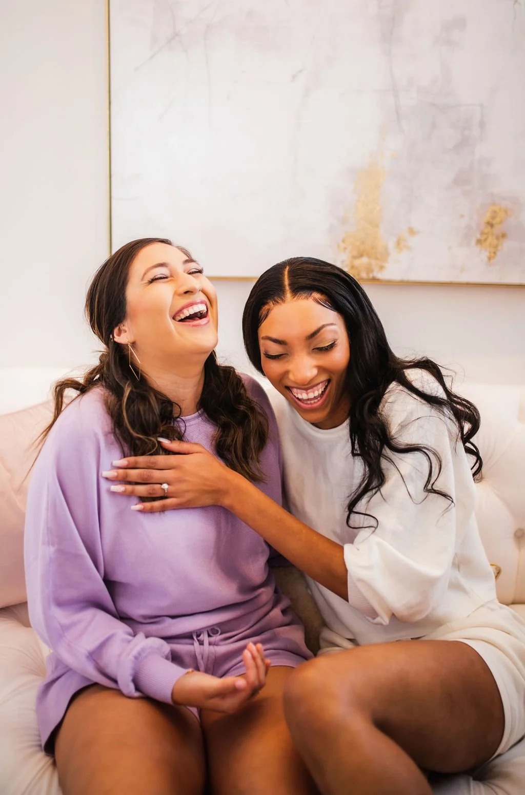 Two women sitting on a couch laughing and smiling, one with her hand on the other's chest.