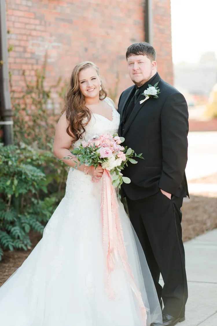 A bride and groom standing outside near a brick wall. The bride is wearing a white wedding dress and holding a bouquet of pink and white flowers with green foliage. The groom is wearing a black suit with a white boutonniere.