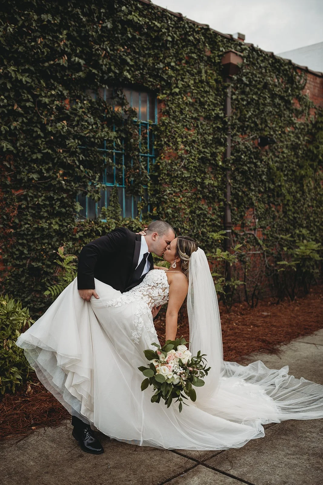 Bride and groom posing in front of the signature green ivy wall at Studio 215 in downtown Fayetteville, NC; a lush urban garden photography backdrop at our industrial warehouse venue near Fort Bragg and Raleigh.