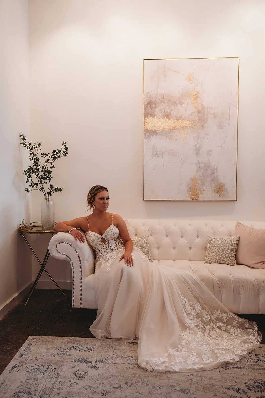 A woman in a white lace wedding dress sitting on a cream tufted sofa in a minimalistic room with white walls, a large abstract painting, a small table with a vase and plant, and decorative pillows.