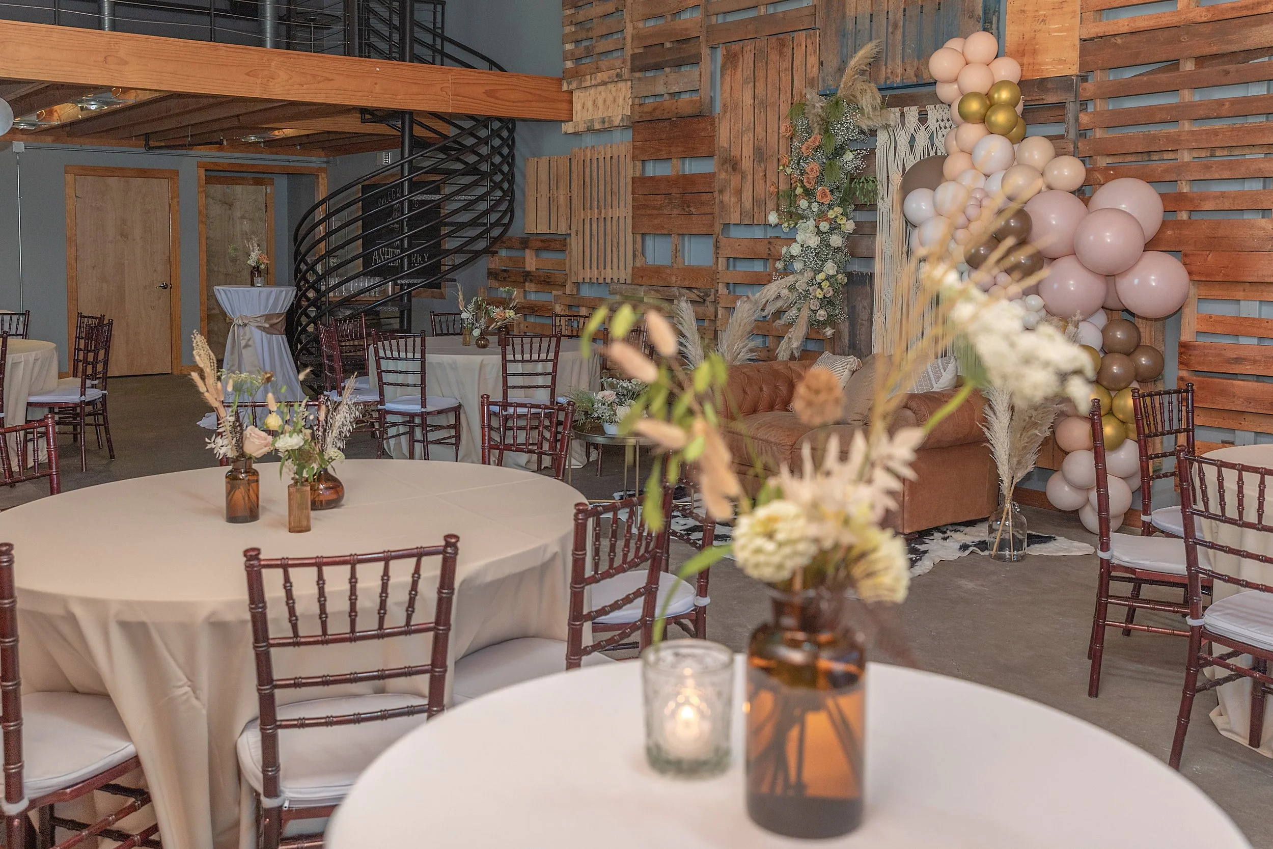 Decorated event space with round tables covered in beige tablecloths, each with small flower arrangements in brown vases; a balloon arch in shades of pink, cream, and gold; wood-paneled and brick wall backdrop; floral arrangements and a brown sofa on one side.