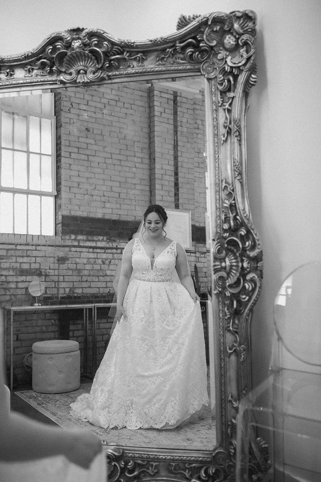 A bride in a lace wedding dress smiling in front of a large ornate mirror.