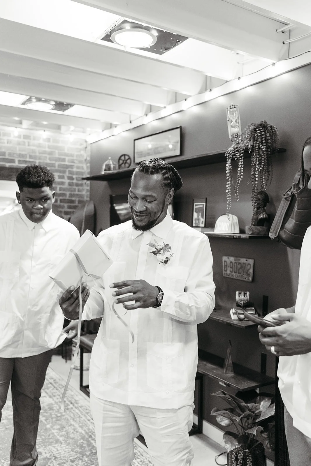 A man in a white suit with a boutonniere is smiling and reading a card, surrounded by two young men also dressed in white, in a decorated indoor space.