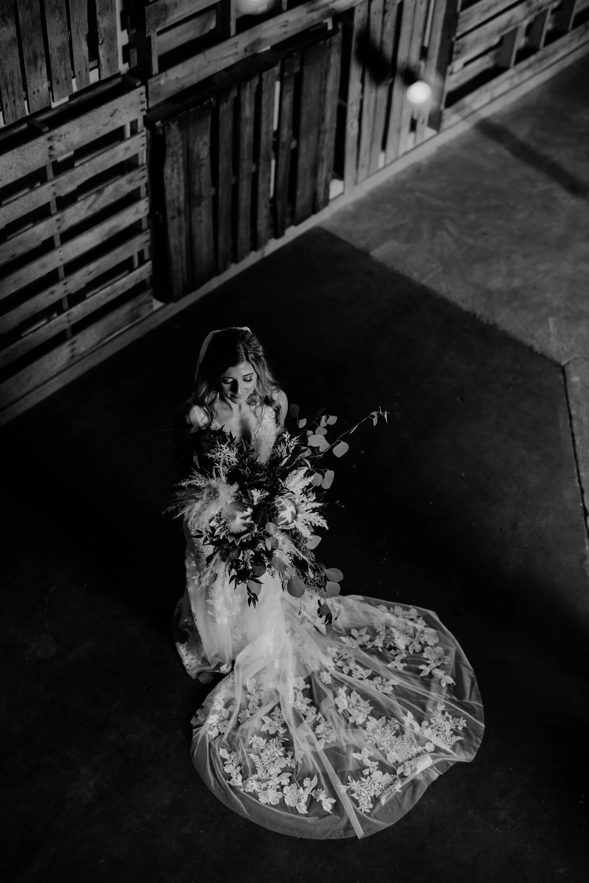 Black and white photo of a bride standing on a dark floor in front of a wooden wall, holding a large bouquet of flowers and greenery, wearing an intricate lace wedding gown with a long floral-embroidered train.