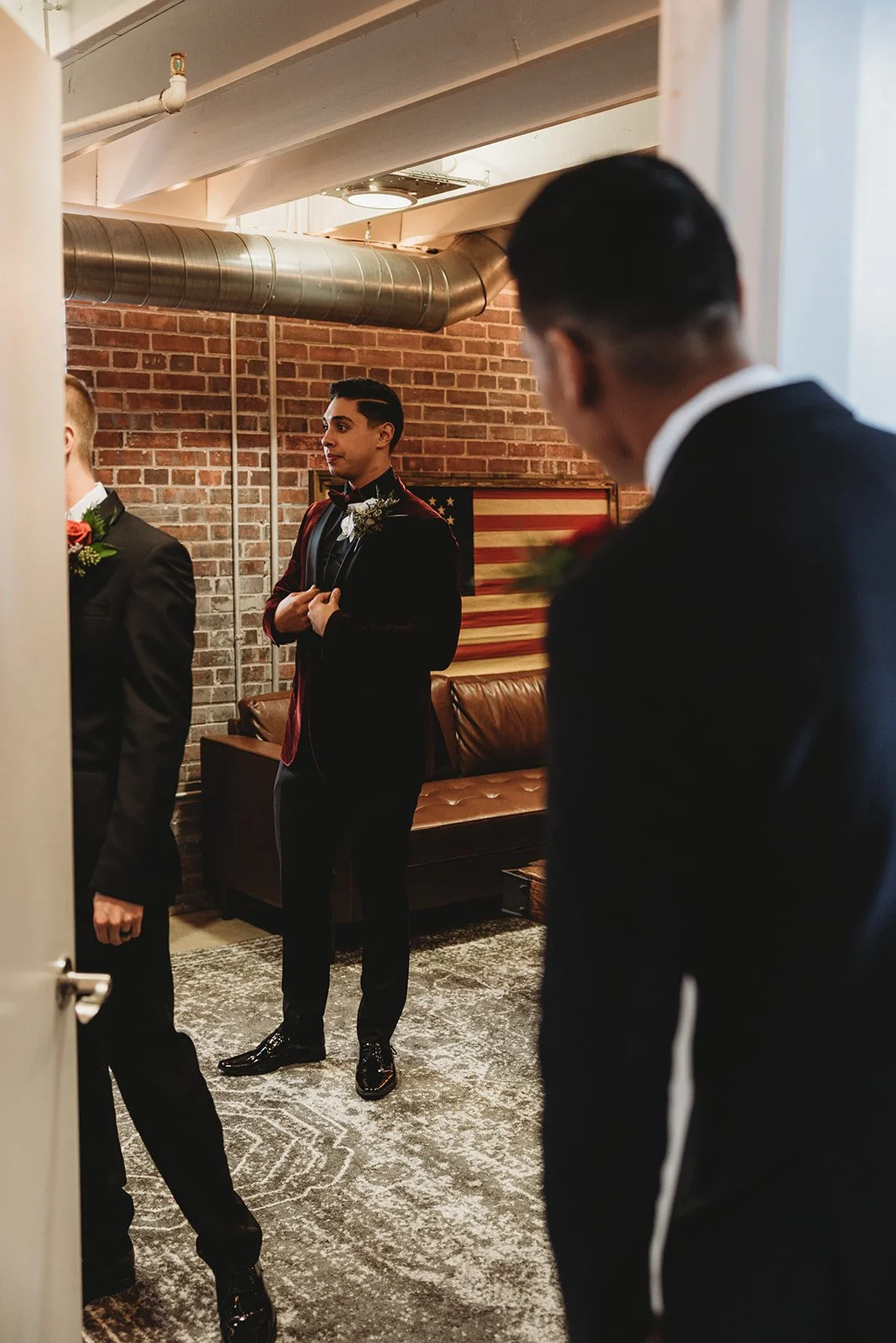 Three men dressed in formal tuxedos standing in a room with brick wall and American flag artwork.