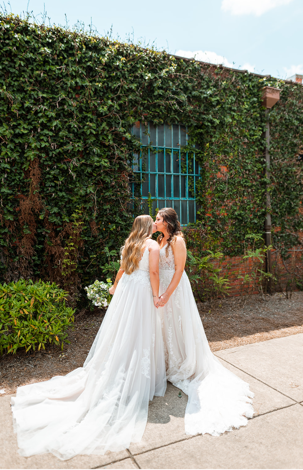 Two women in wedding dresses holding hands and sharing a kiss outdoors, with a green ivy-covered brick wall and a blue window behind them.