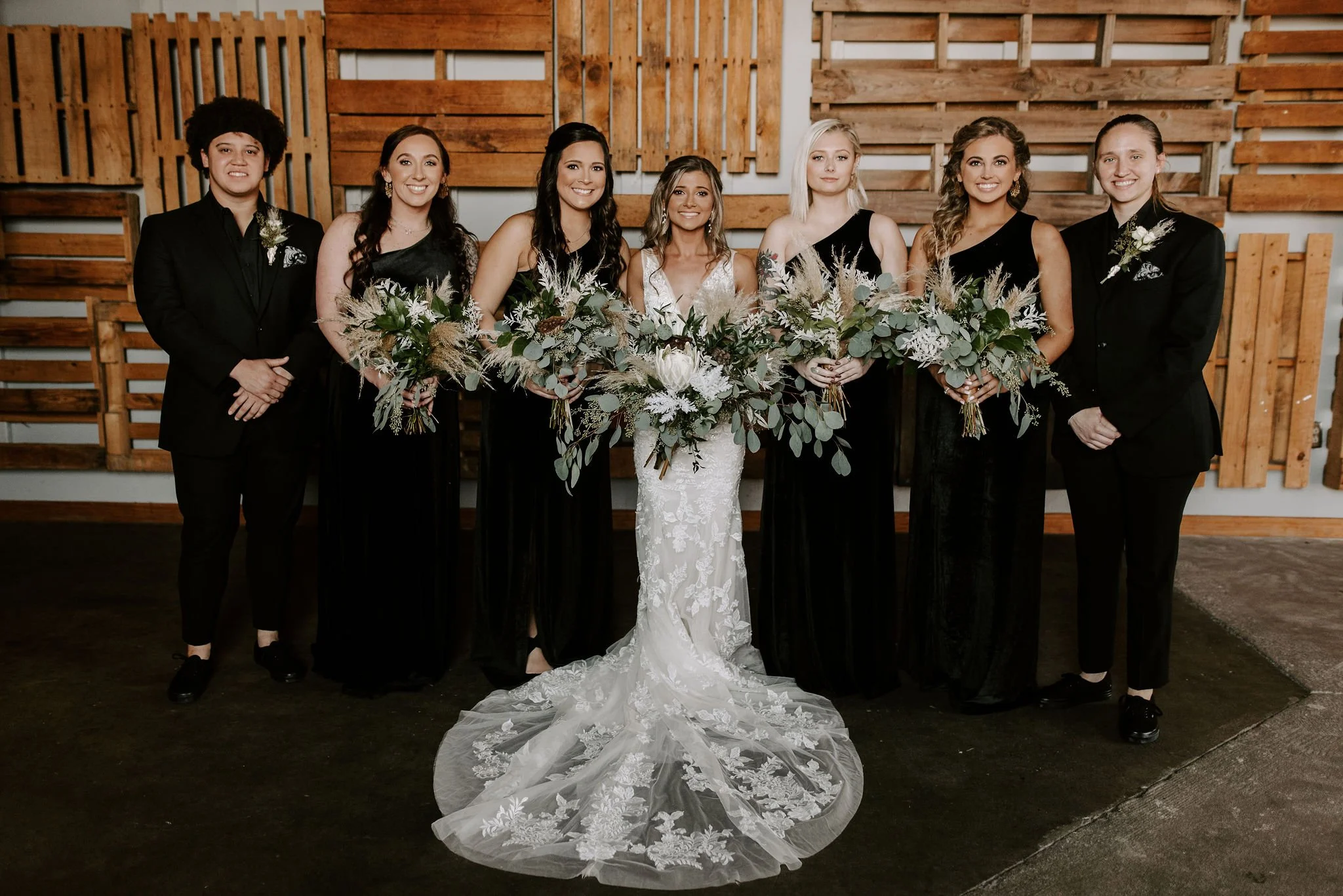 A group of seven women at a wedding, including the bride in a white lace gown and six bridesmaids in black dresses, standing in front of a wooden wall, holding bouquets, smiling for the camera.