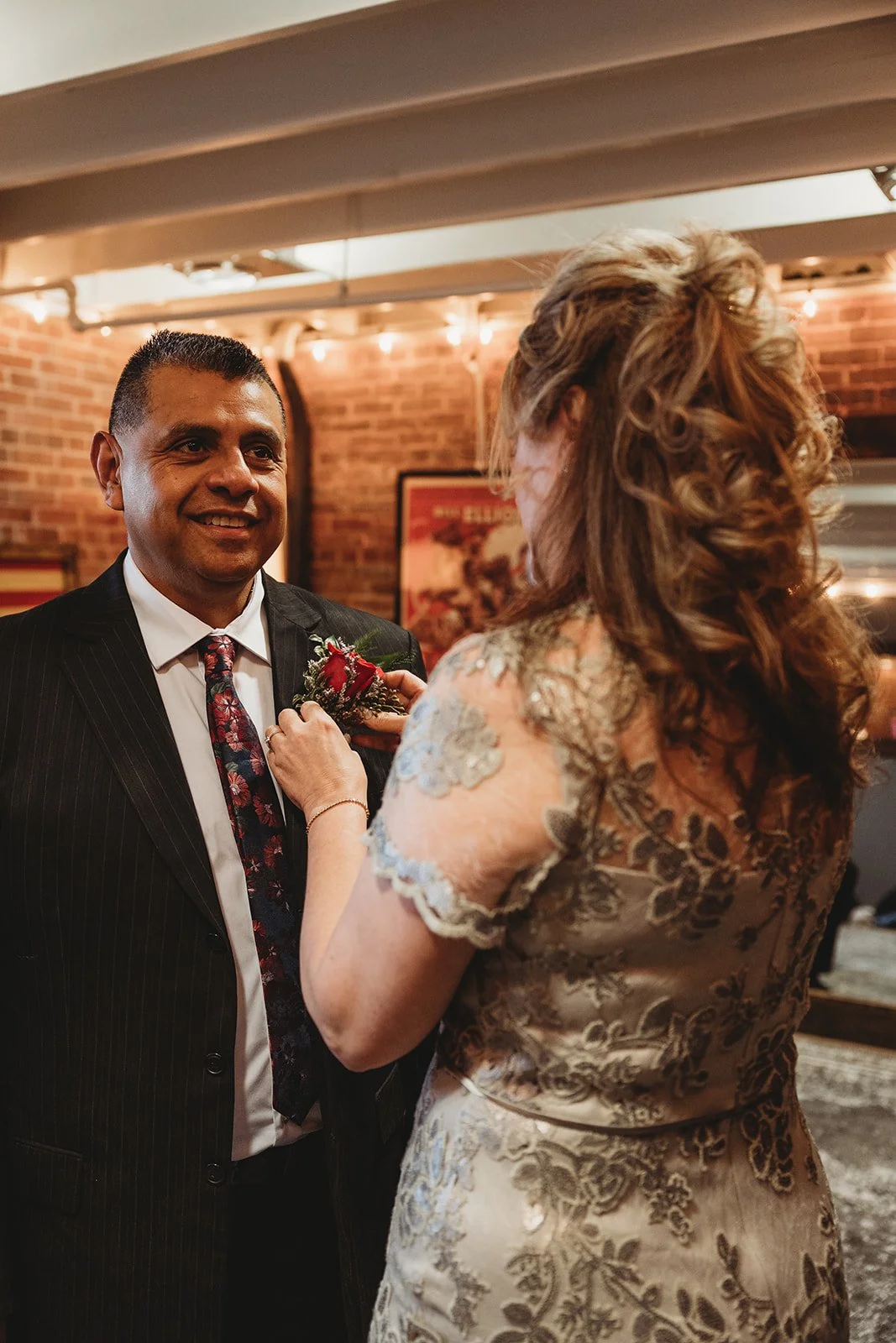 Woman pinning a boutonniere on a smiling man in a suit at an indoor event with brick walls and warm lighting.